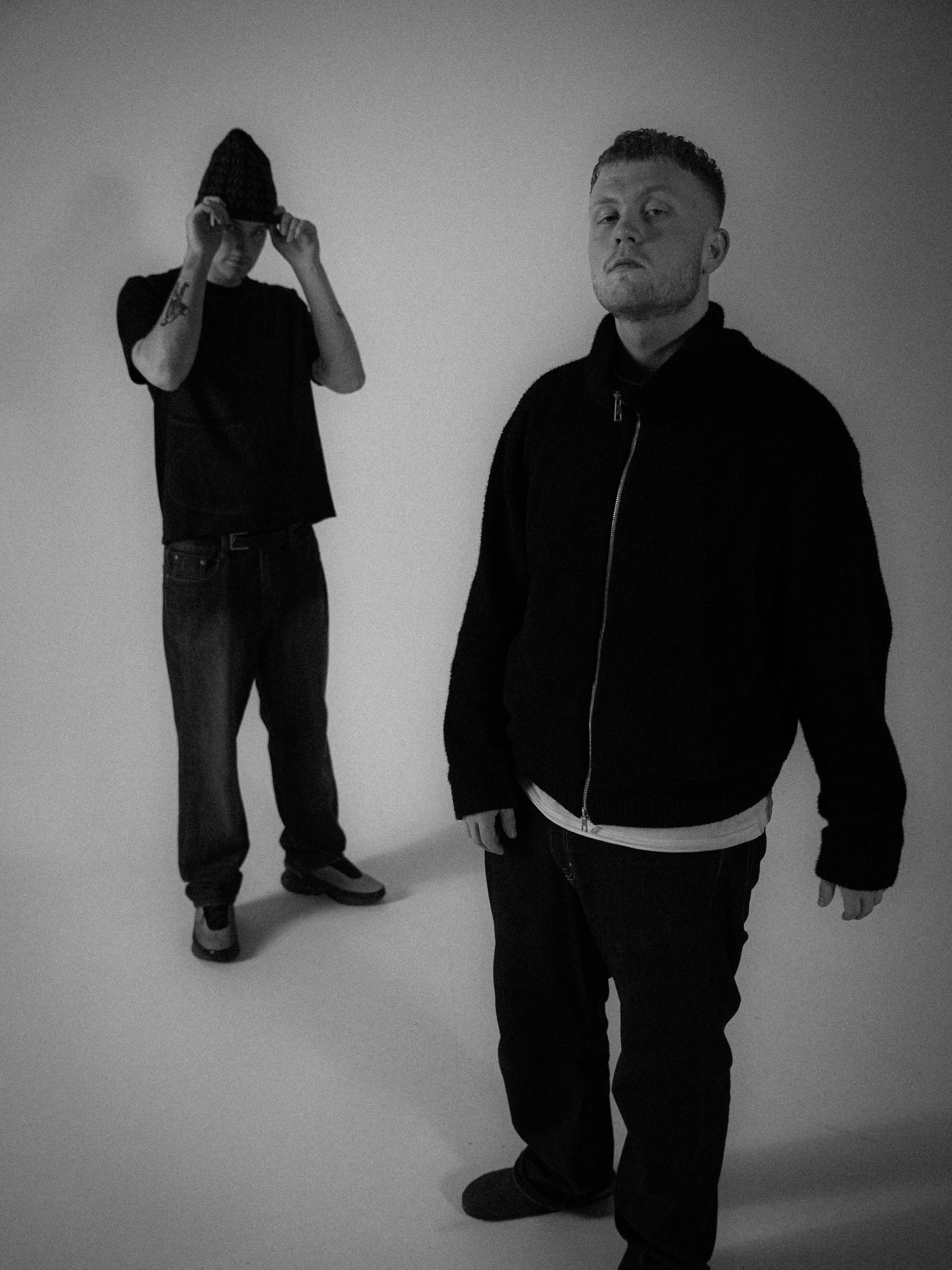 Two young men with casual clothing pose against a plain background. One is forward-facing with a serious expression, and the other is adjusting his hoodie while looking downward.