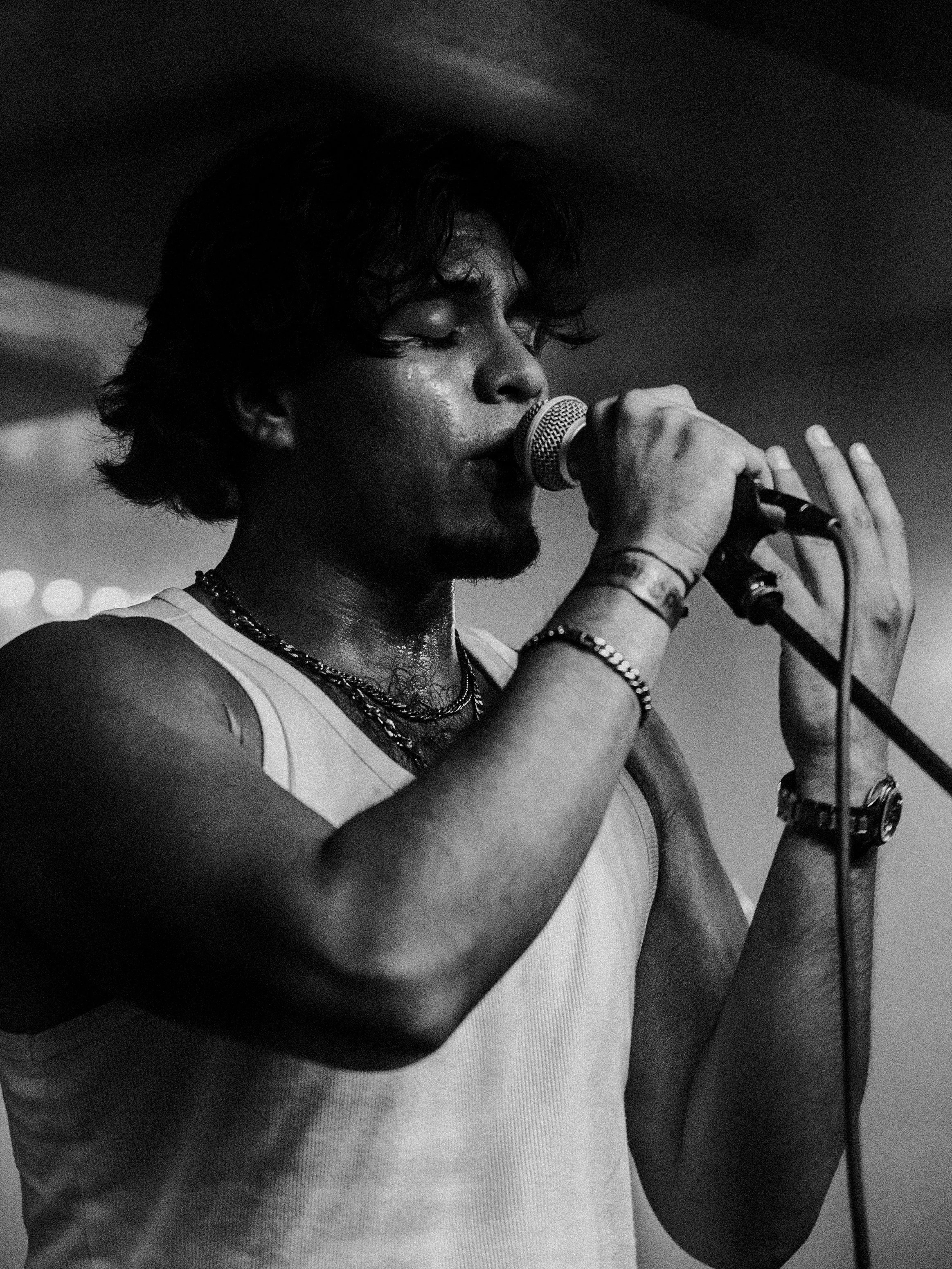 A black and white photo of a young man singing into a microphone with his eyes closed, wearing a sleeveless shirt and multiple bracelets.