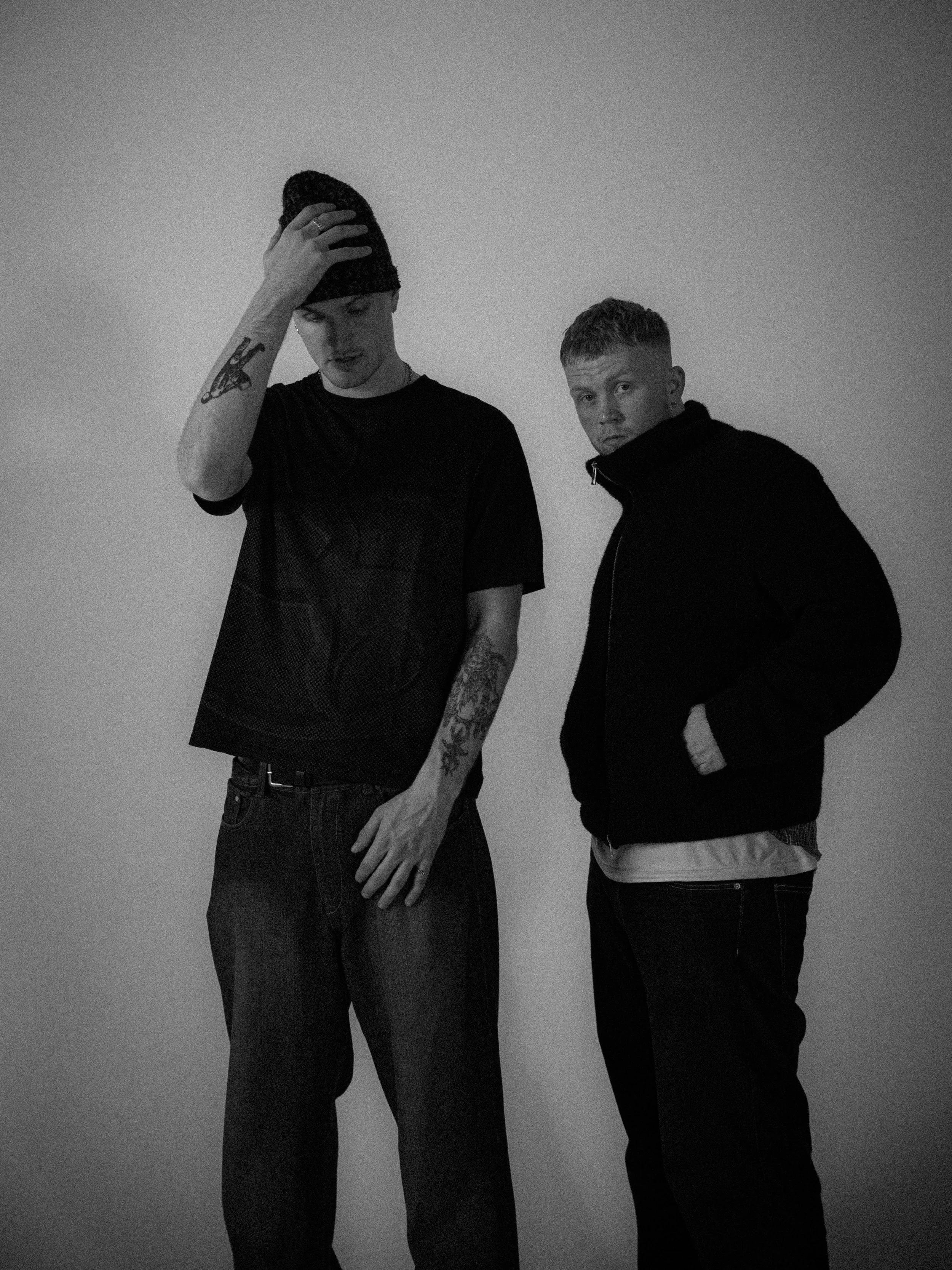 Two young men stand against a plain wall, one with hand on head and tattoos on arm, the other with hands in jacket pocket, both looking at the camera in a black and white photo.