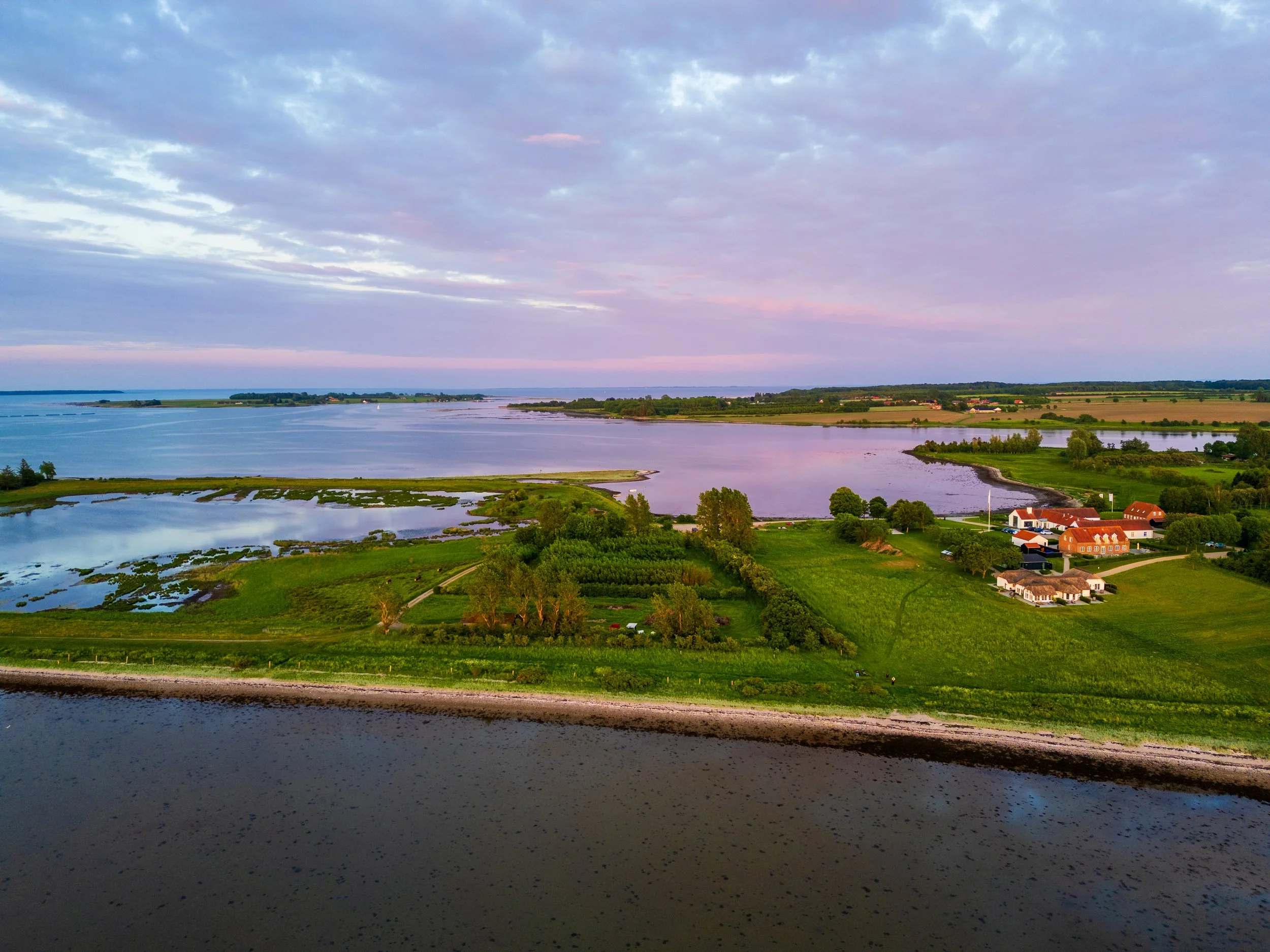 Aerial view of a landscape featuring a body of water, green fields, trees, and small buildings at sunset or sunrise, with a partly cloudy sky.