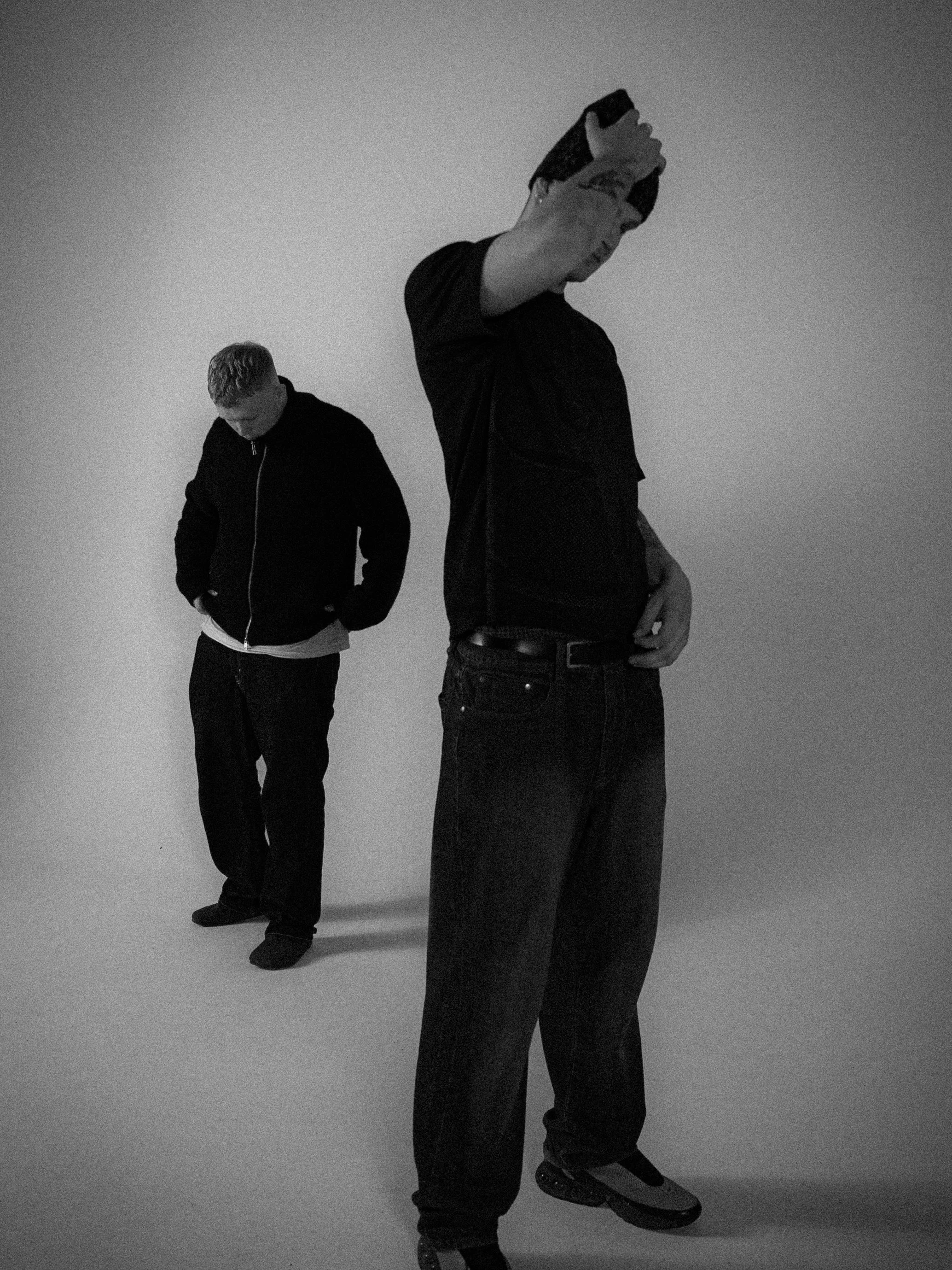 Two young men standing in a plain white room, one in the foreground with a camera and the other in the background with hands in pockets.