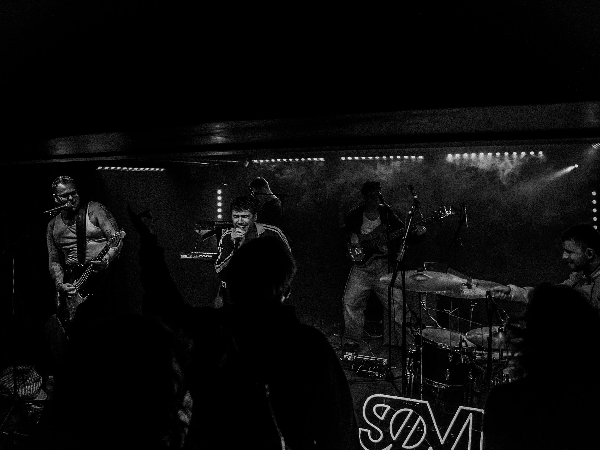 A live band performs on stage in a dimly lit venue, with a singer and guitarists, and audience members enjoying the performance.