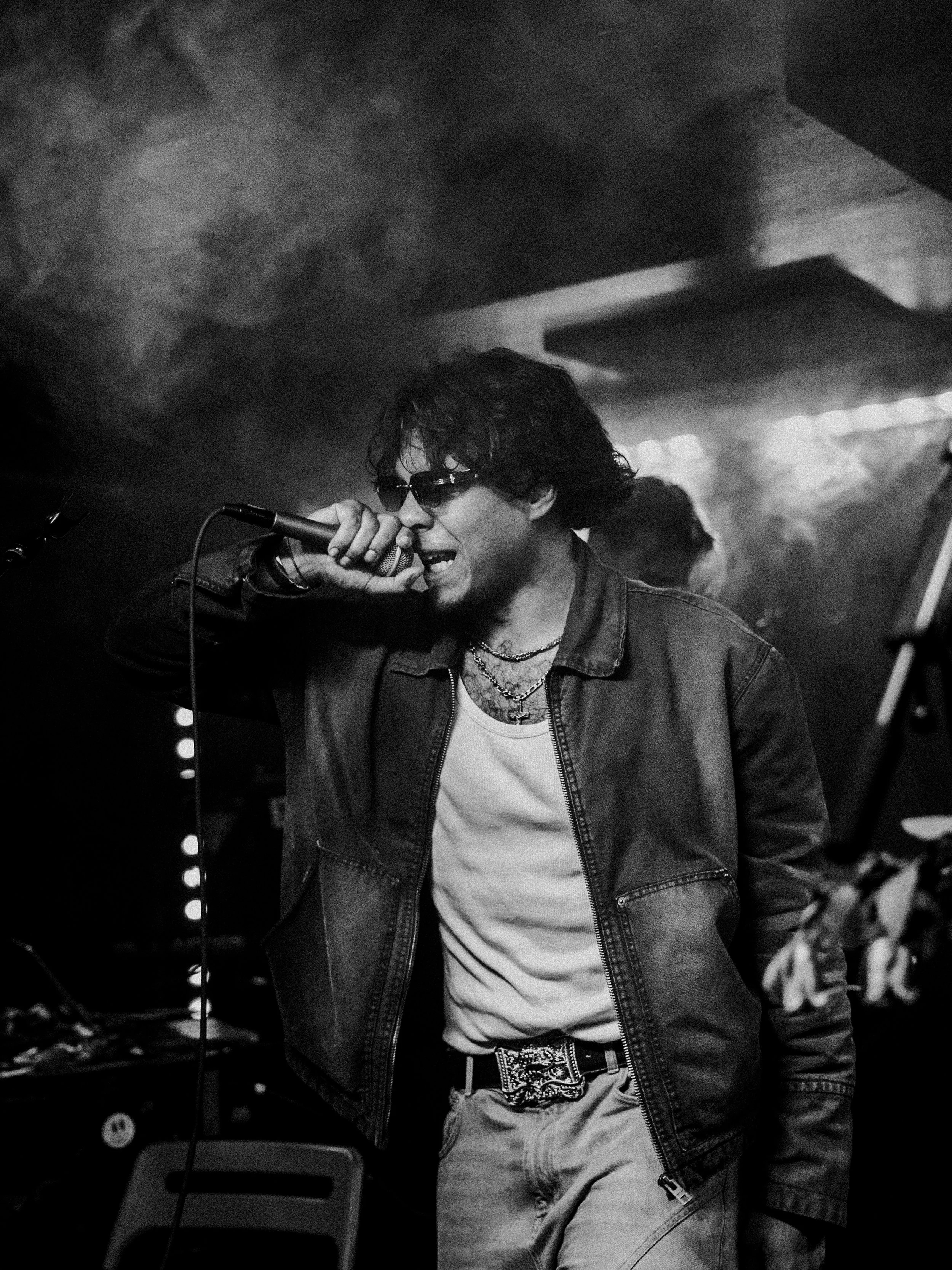 A man singing into a microphone on stage, wearing sunglasses, a denim jacket, and layered necklaces, in a dimly lit music venue.