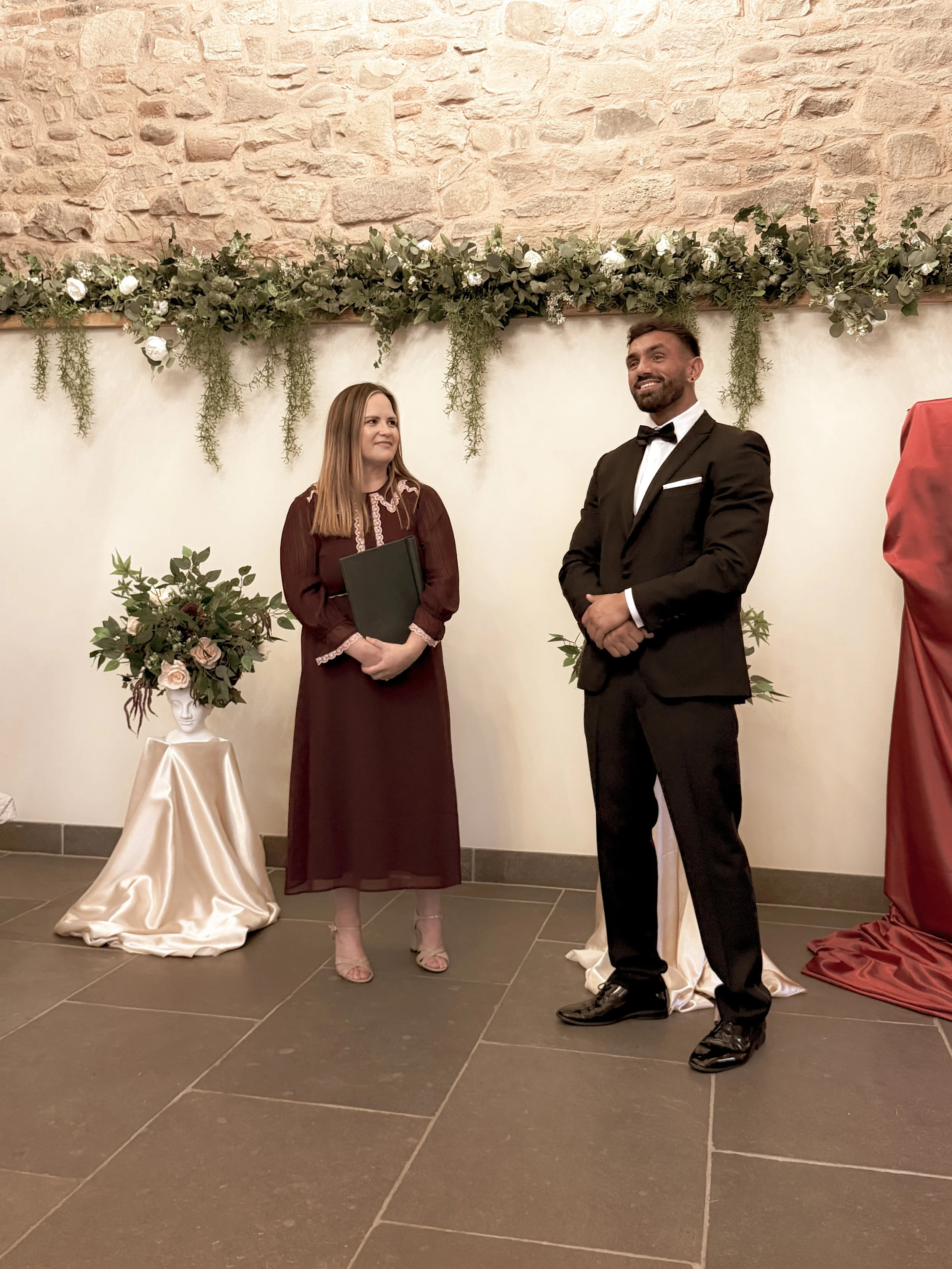 Emily Ayres in a maroon dress stands next to a groom in a black tuxedo at a wedding. Behind them is an elegant white wall decorated with a lush green and white floral arrangement and hanging greenery.