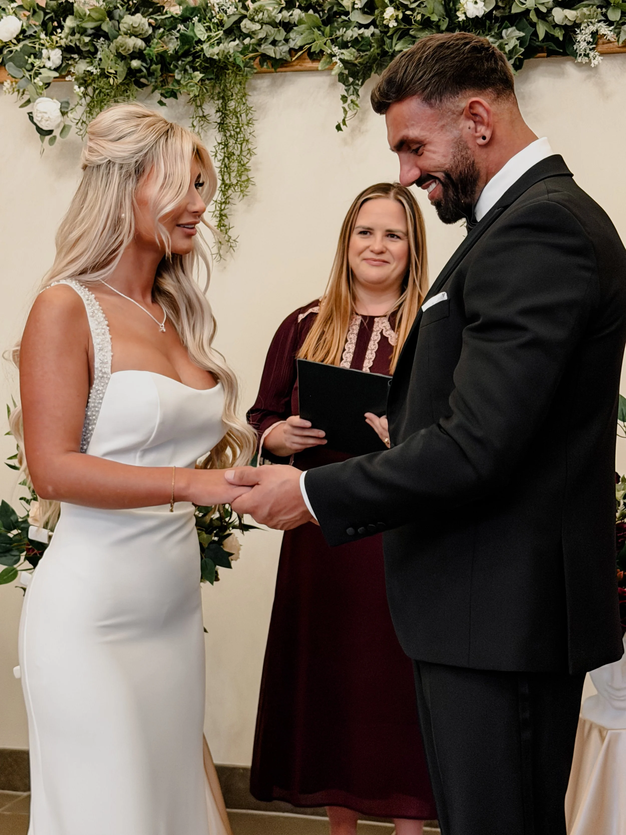 A bride and groom holding hands during a wedding ceremony, with Emily Ayres standing behind them, decorated with floral arrangements and greenery.