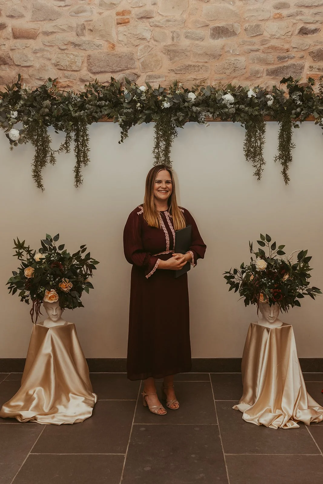 Emily Ayres in a burgundy dress with pink lace details stands smiling at a wedding ceremony in front of a decorative floral backdrop, with two mannequins topped with floral arrangements on either side.