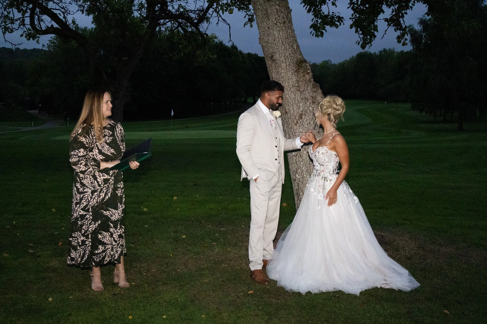 A wedding ceremony outdoors at dusk, with a bride and groom exchanging rings beside a large tree, Emily Ayres holding a folder with their ceremony script, standing nearby, in a green park setting.