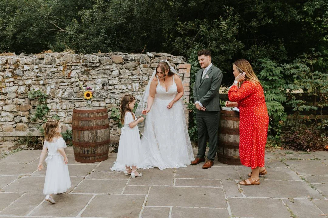 Wedding scene outdoors with a bride and groom, two young flower girls, and Emily Ayres, South Wales Celebrant is helping the young girls with the wedding rings. There are two wooden barrels and a stone wall with greenery in the background.