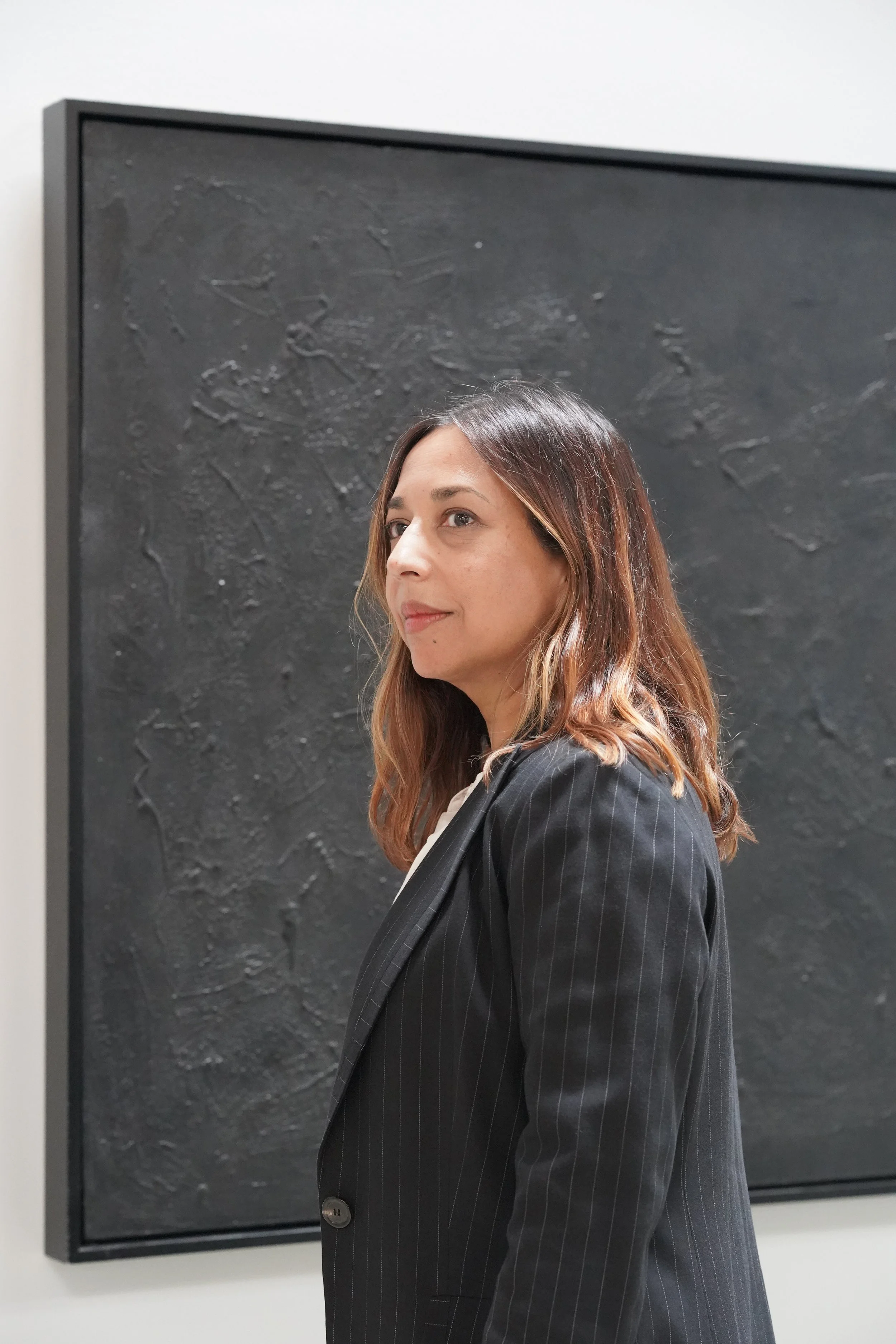 A woman with shoulder-length brown hair stands in front of a textured black artwork at an art gallery.