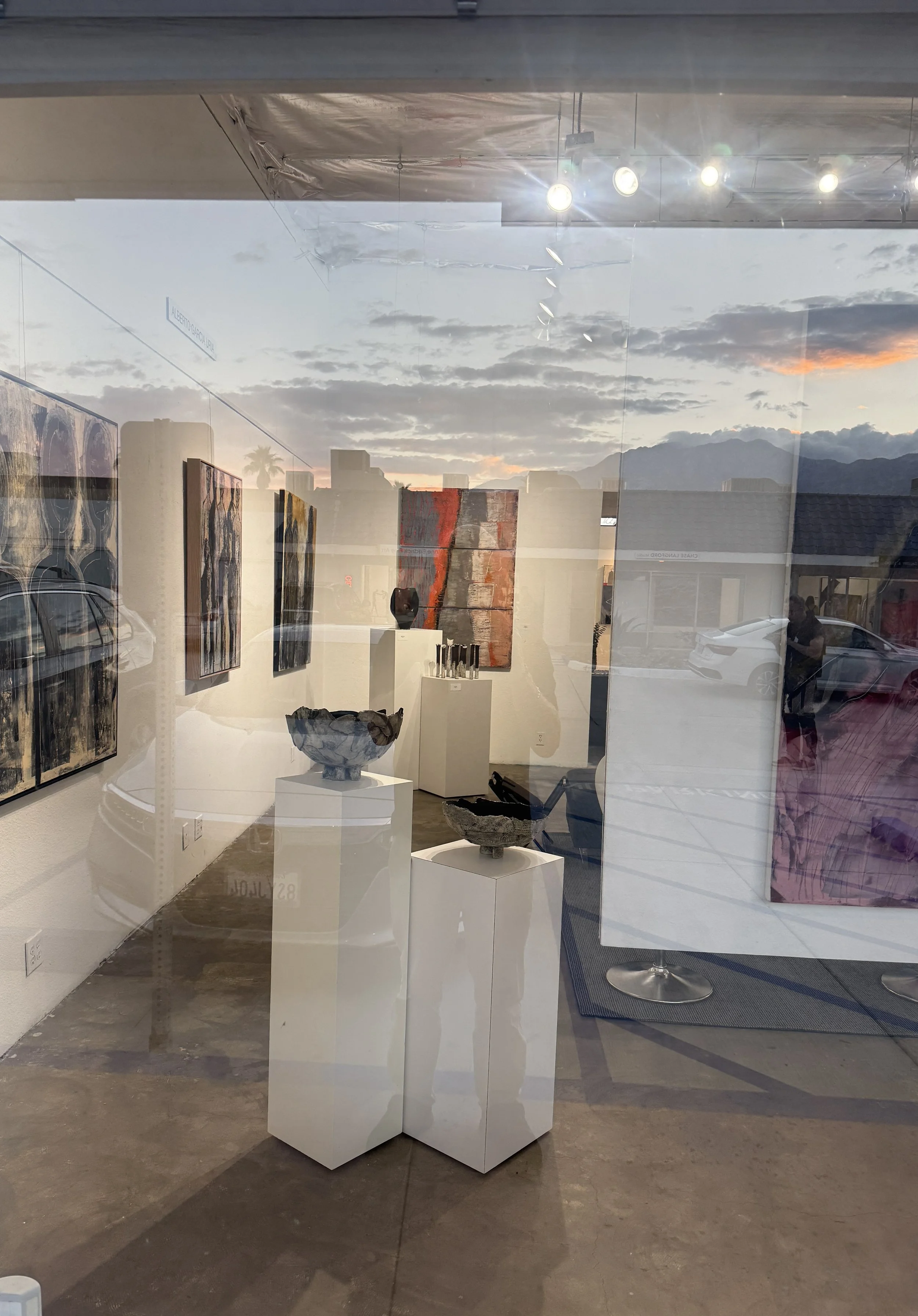 View of an art gallery with paintings on the wall and sculptures on pedestals, seen through glass when sunset sky and clouds are visible outside.