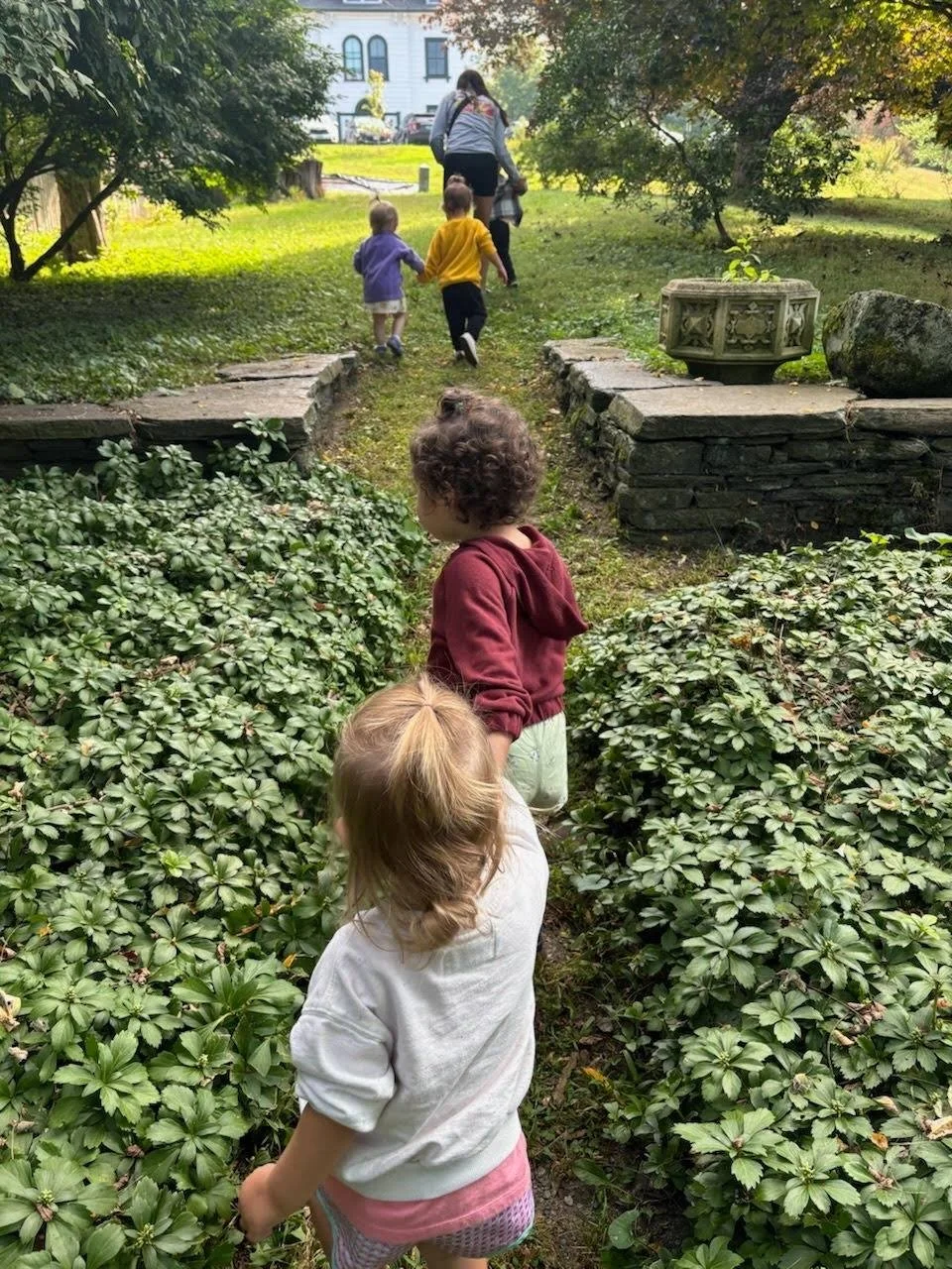 Children walking along a garden path with an adult in a park-like setting surrounded by trees and shrubs.