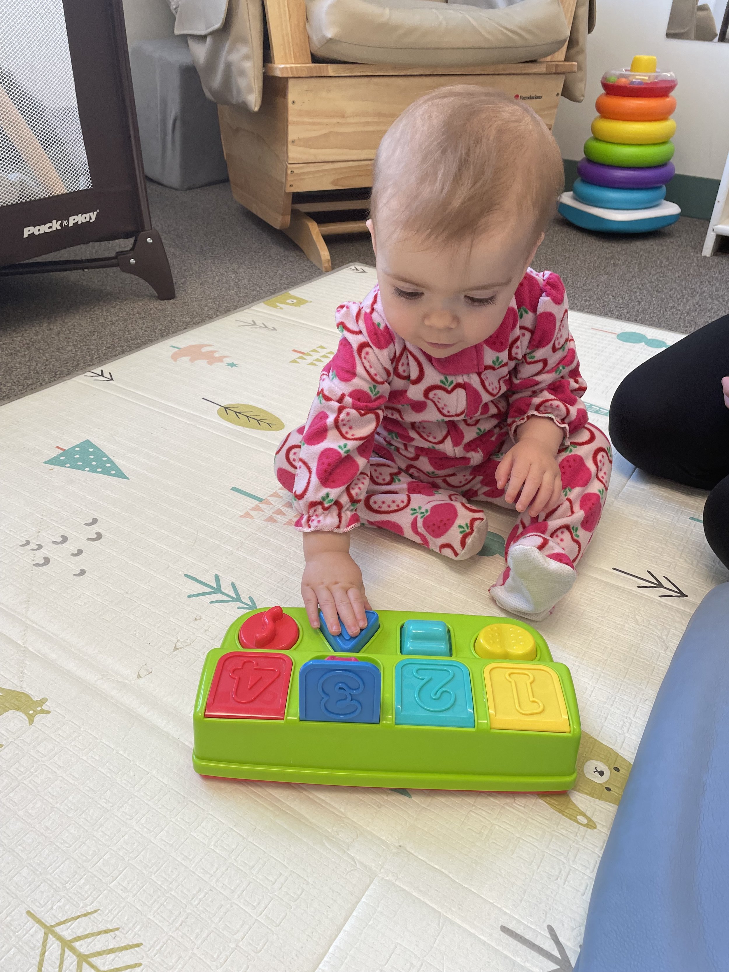A toddler girl in a strawberry-patterned pajamas playing with a colorful shape-sorting toy on a patterned mat in a playroom.