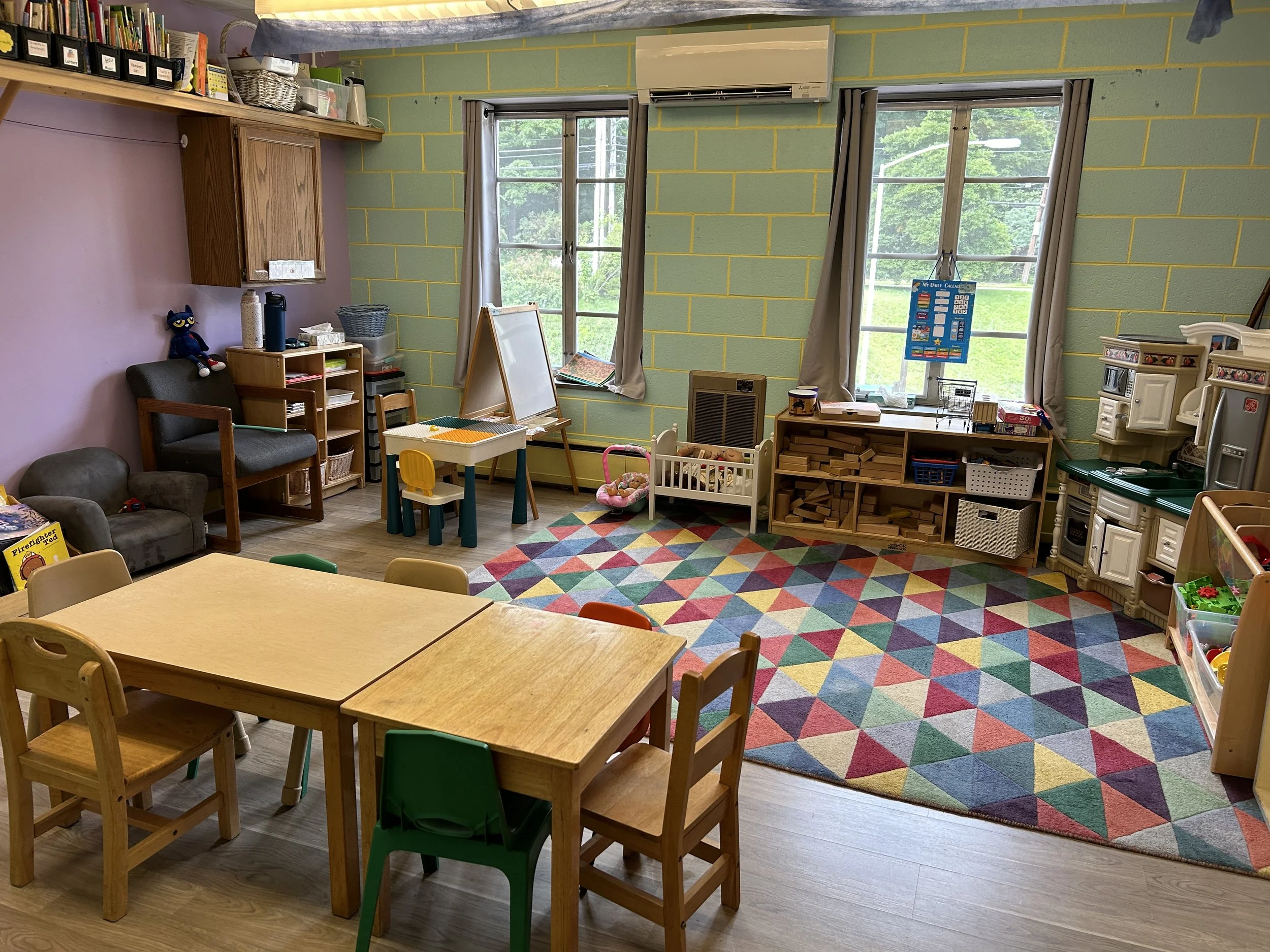 A preschool classroom with wooden tables and chairs, a colorful geometric rug, toy kitchen, and various educational materials and stuffed animals, with windows showing greenery outside.