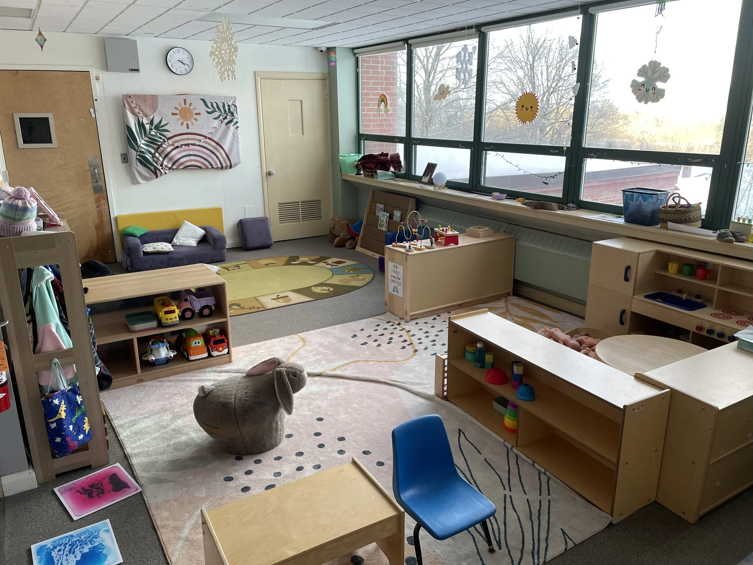 A colorful preschool classroom with toys, furniture, and decorations. There is a large window with snow outside, a clock on the wall, and various art projects hanging. The room has a cozy and organized setup for children to play and learn.