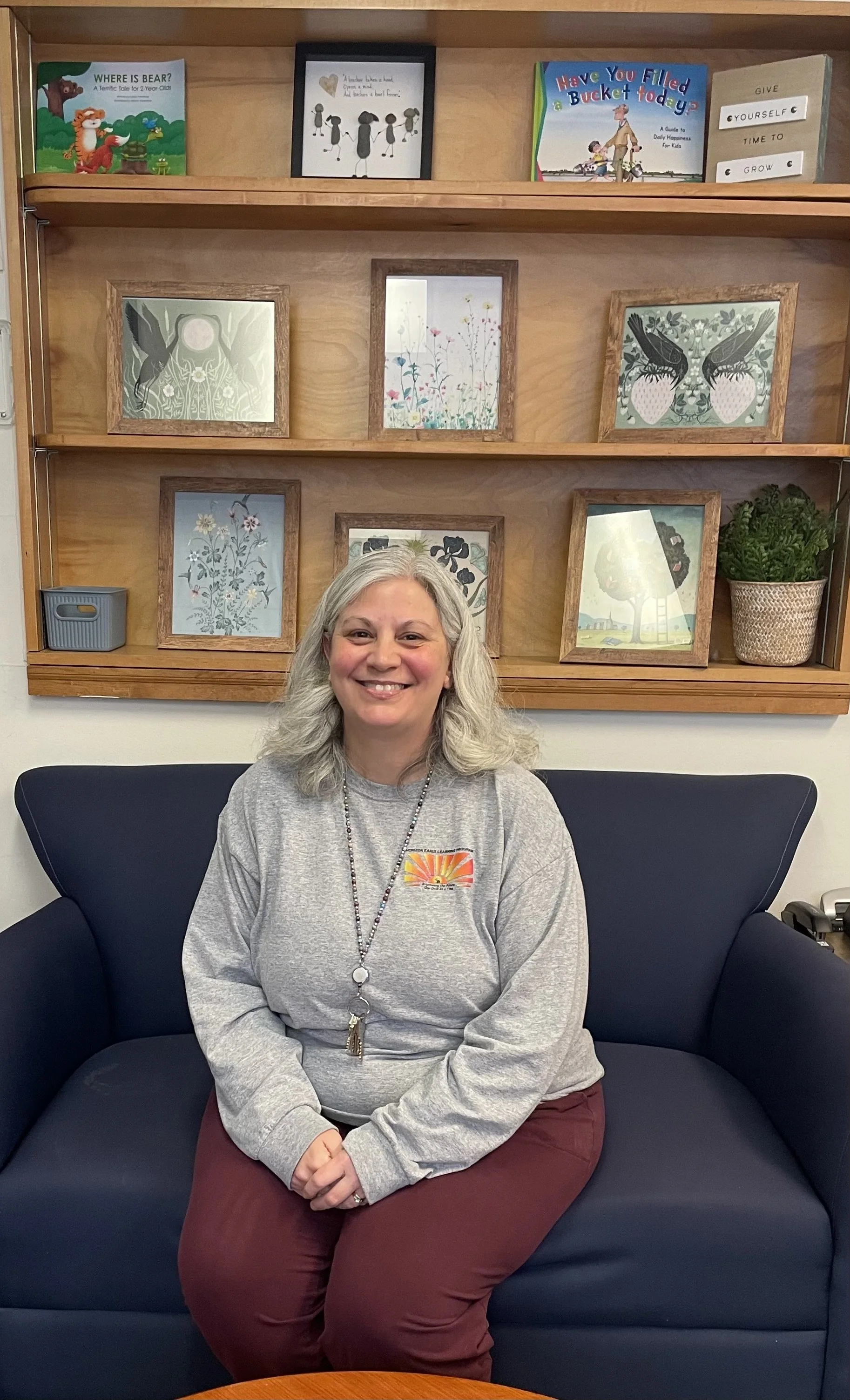 A woman with gray hair sitting on a dark blue sofa in a room with a wooden shelf behind her, featuring framed artwork and a potted plant.