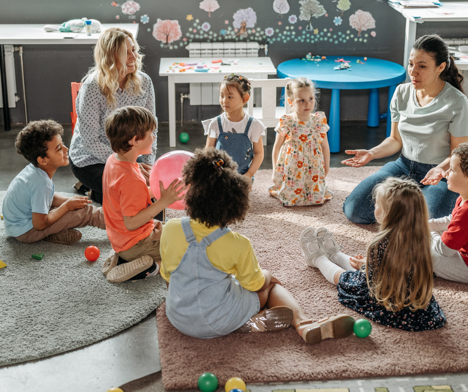 Two adult women and children sitting on colorful rugs in a classroom with a dark wall decorated with tree and flower art, engaging in discussion or activity.