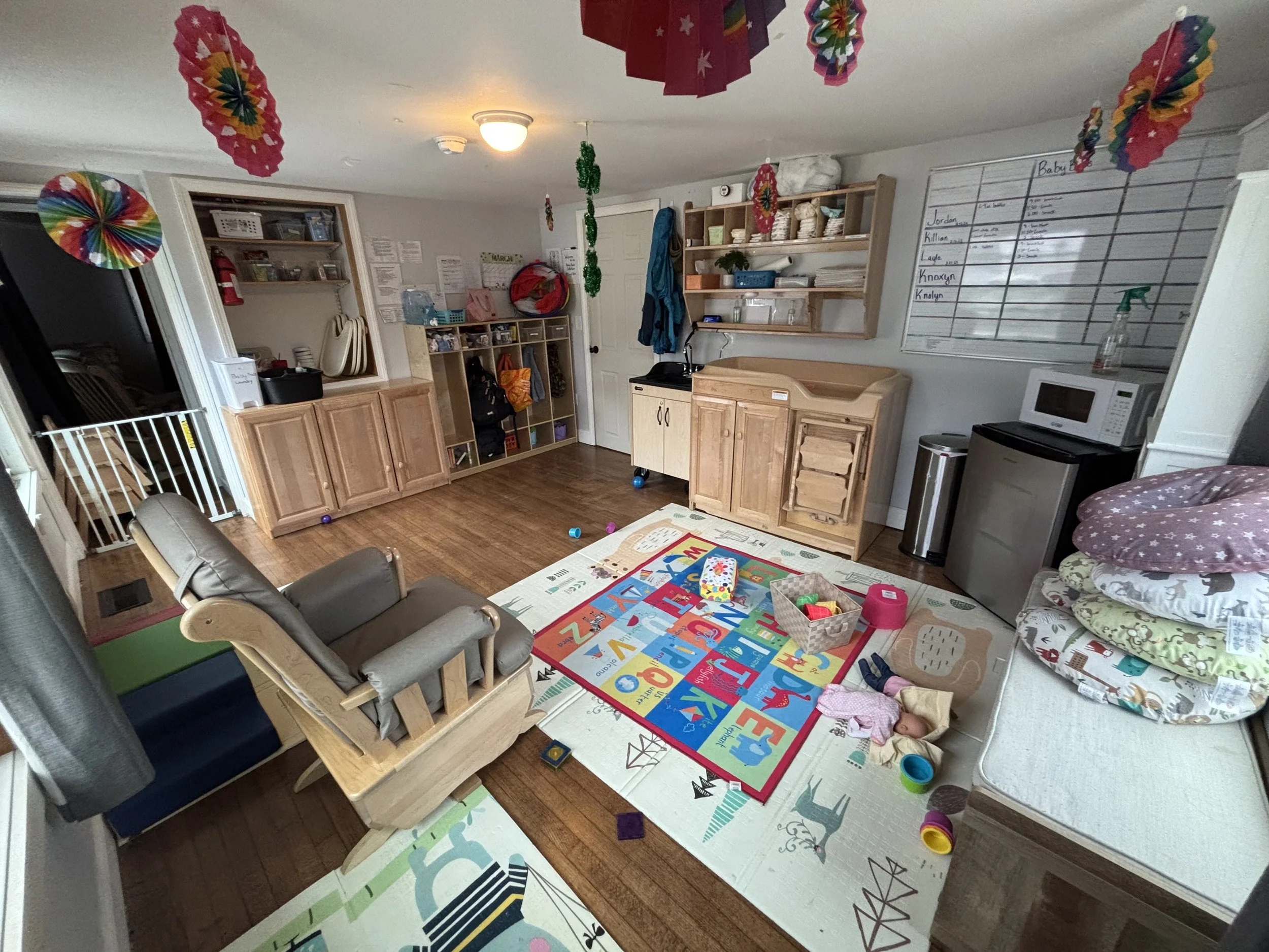 A cluttered playroom with colorful decorations, a children's rug, a doll on the floor, and various toys and furniture.