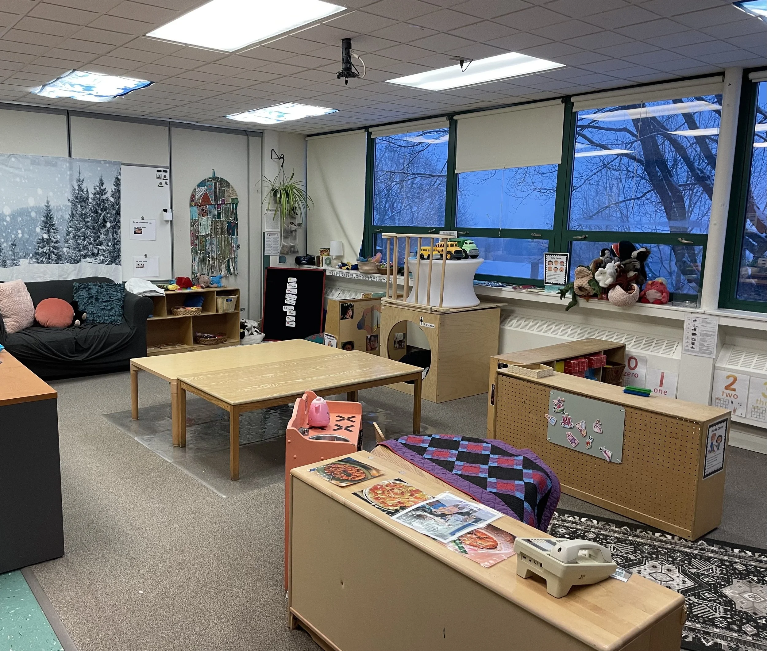 A classroom with large windows showing a snowy outdoor scene, decorated with toys, craft supplies, and educational materials, with a sofa, tables, and shelves.
