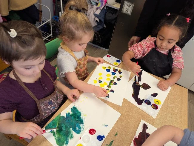 Four young girls at a table painting with various colors on paper in an art classroom or activity room.