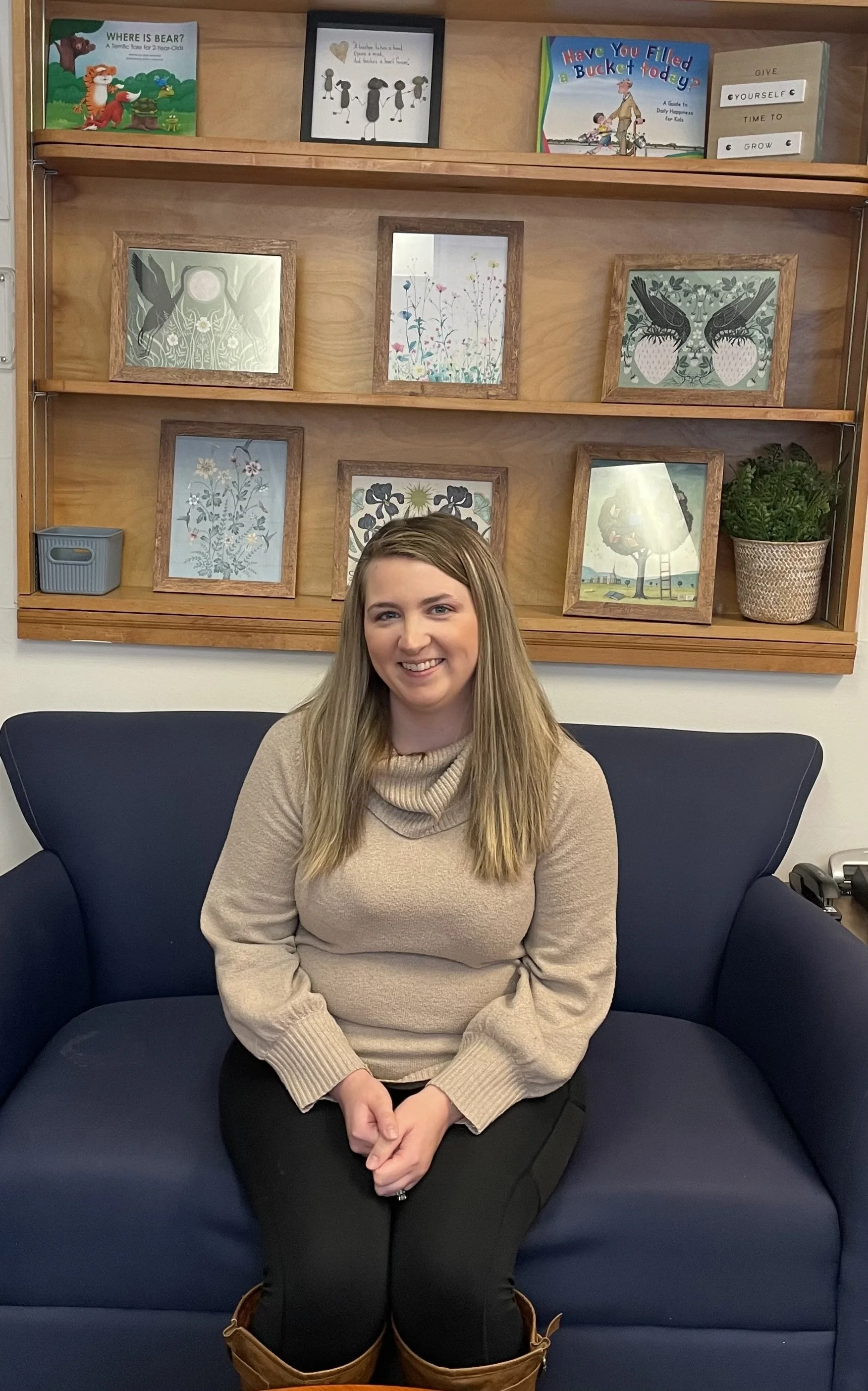A young woman sitting on a dark blue couch in front of a wooden wall shelf with framed artwork and books.