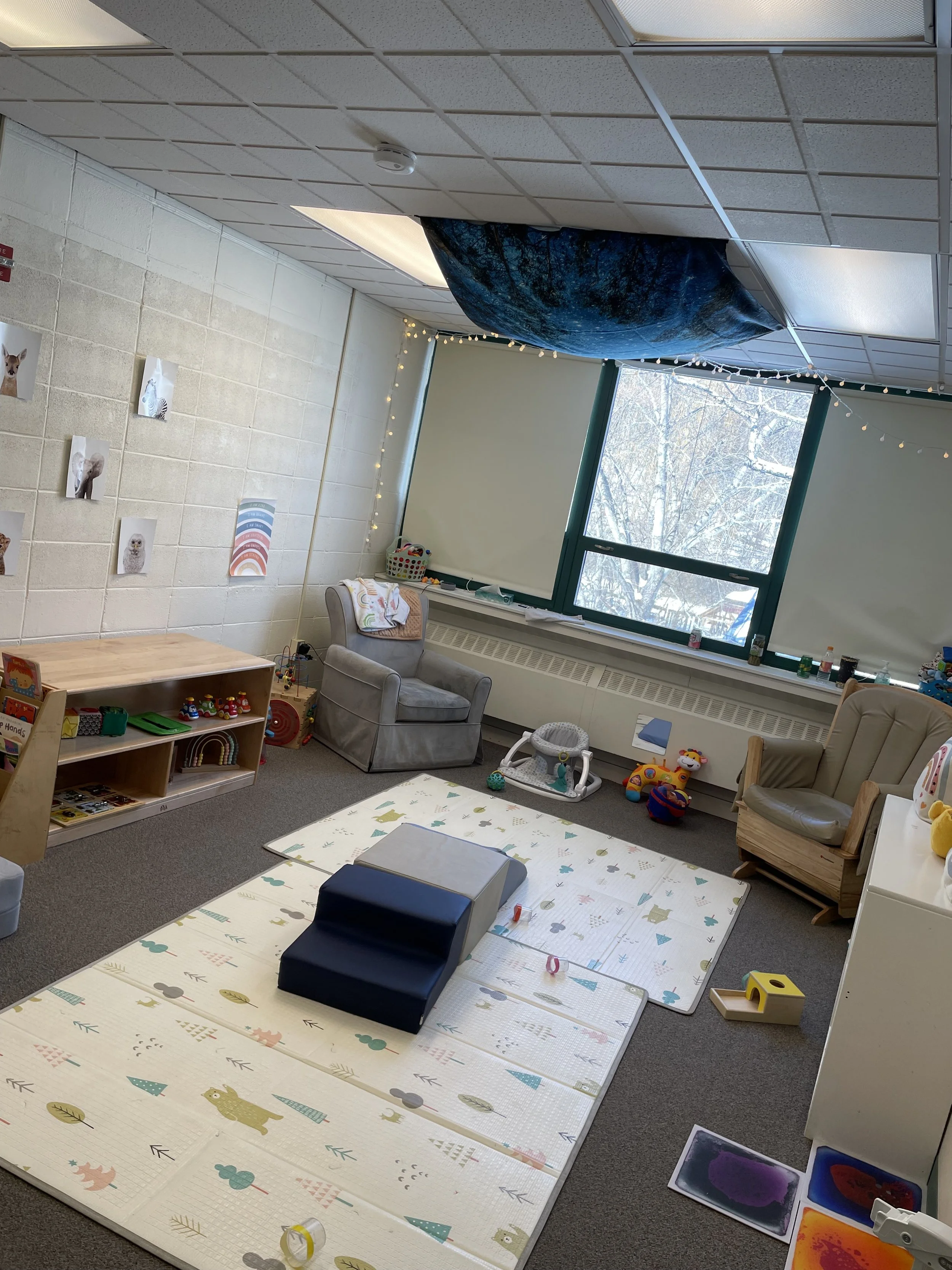 Indoor children's playroom with foam mats, toys, chairs, and window with snow outside.