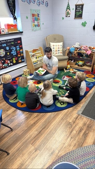 A man reading a book to six young children sitting on a colorful alphabet rug in a classroom.