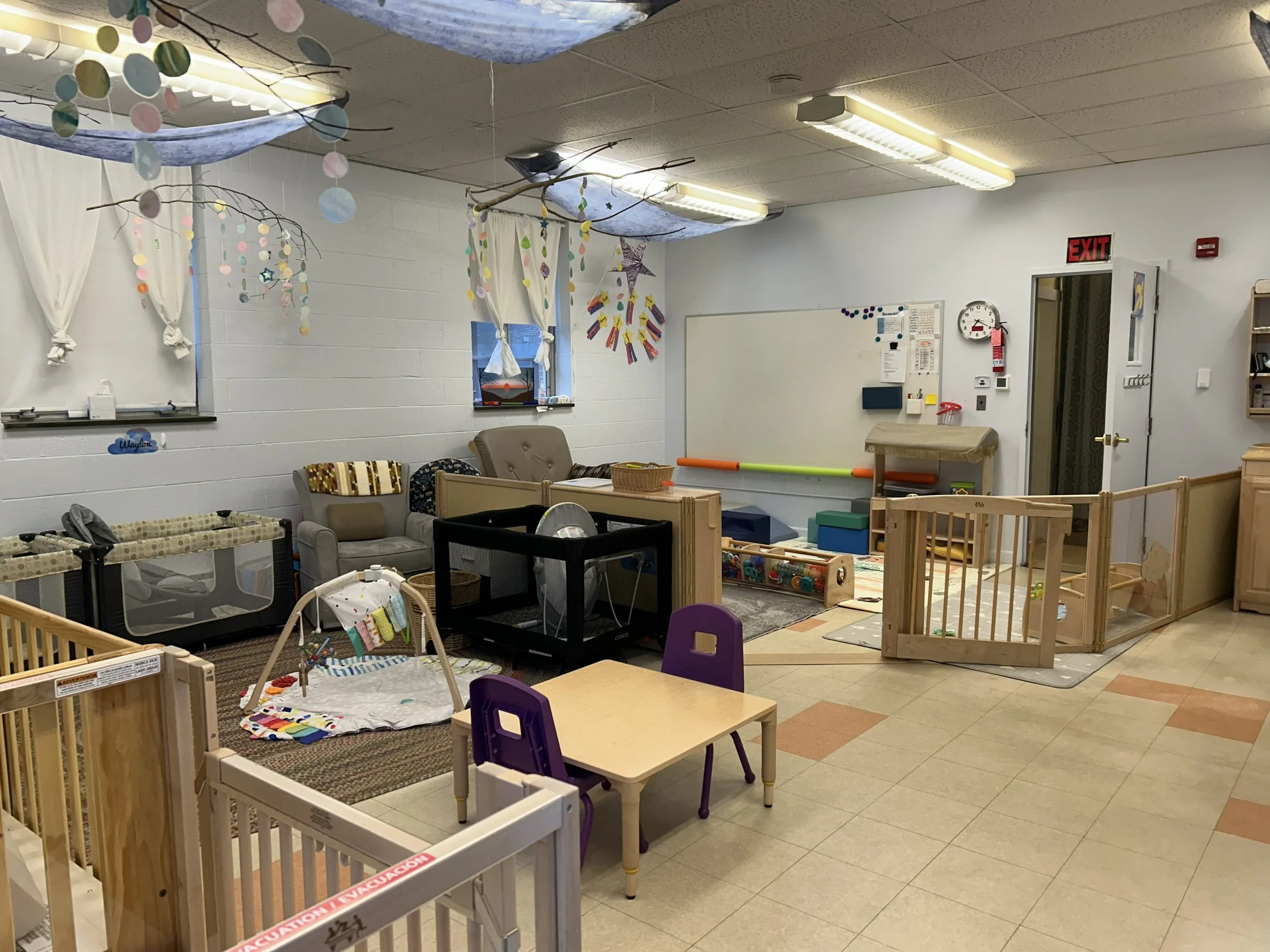 Indoor daycare or playroom with small wooden and fabric play fences, a small table with purple chairs, seating area with sofa, toys and play mats, white walls with windows, and ceiling decorated with hanging paper circles and fabric.