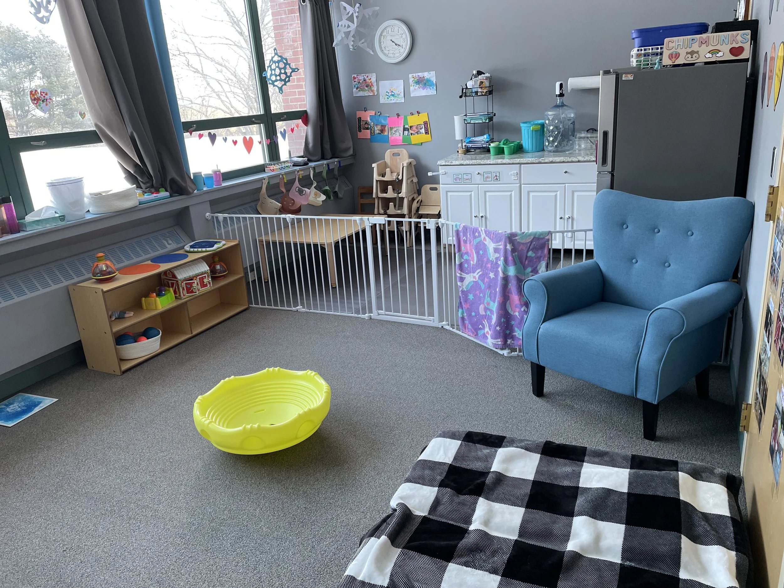 A colorful playroom with a large window, blue armchair, yellow plastic kiddie pool, black and white checkered blanket, and a white safety gate. The room has toys, artwork on the walls, and a white cabinet with a water dispenser on top.
