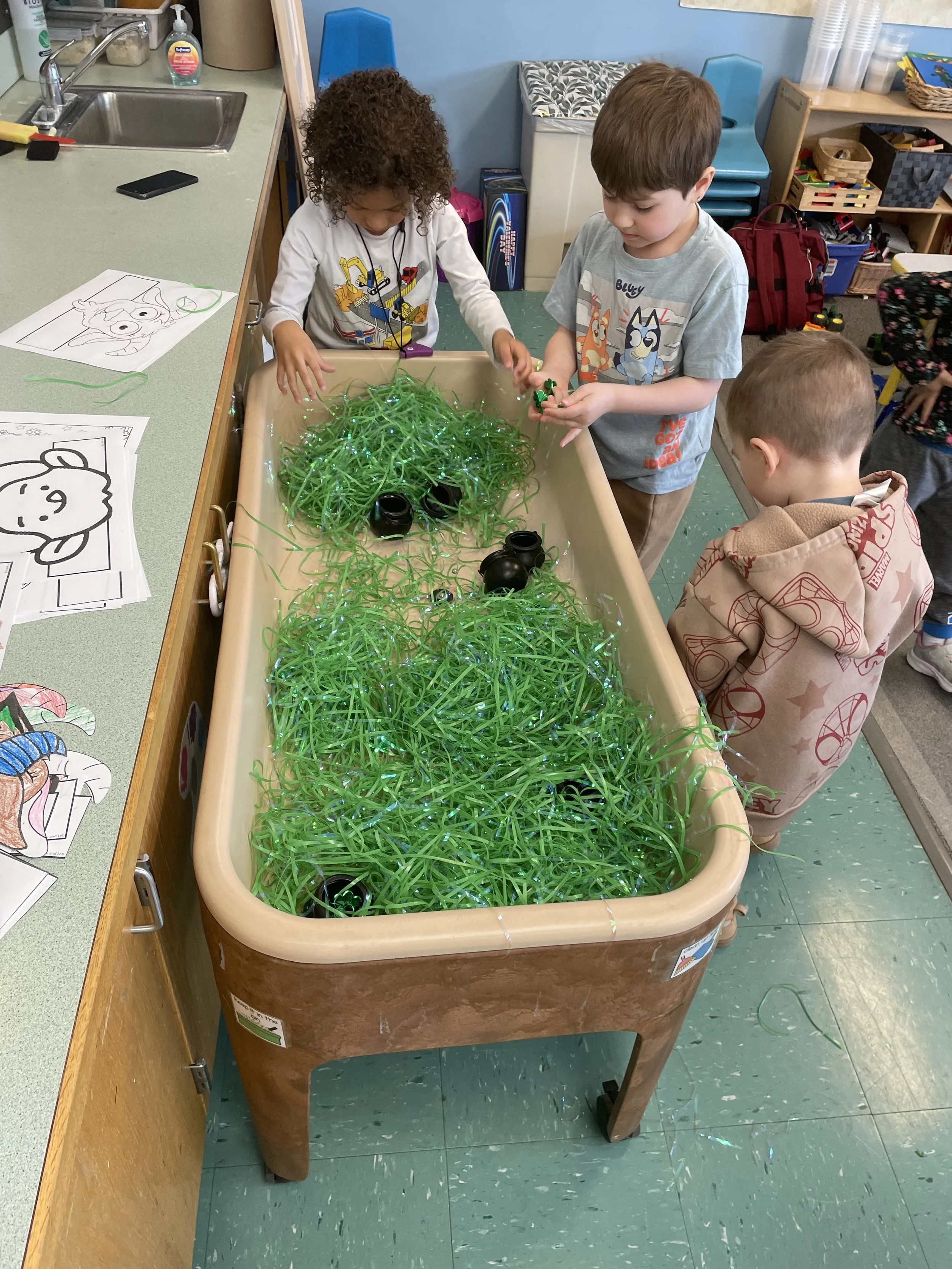 Three children playing with green plastic grass and small black pots in a classroom.