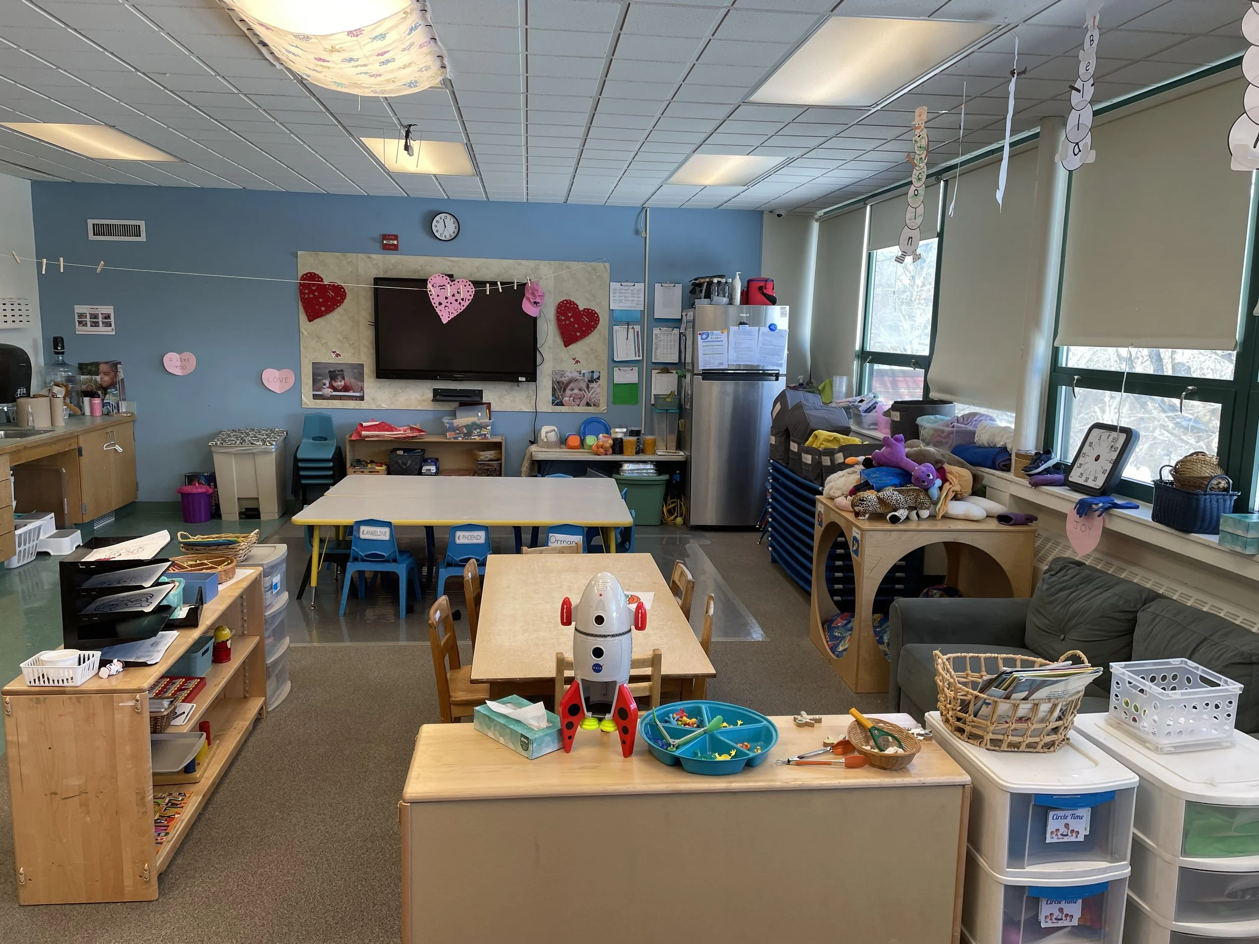 A classroom decorated with valentine's hearts and paper hearts hanging from the ceiling and wall. There are multiple tables, a couch, toys, and various supplies.