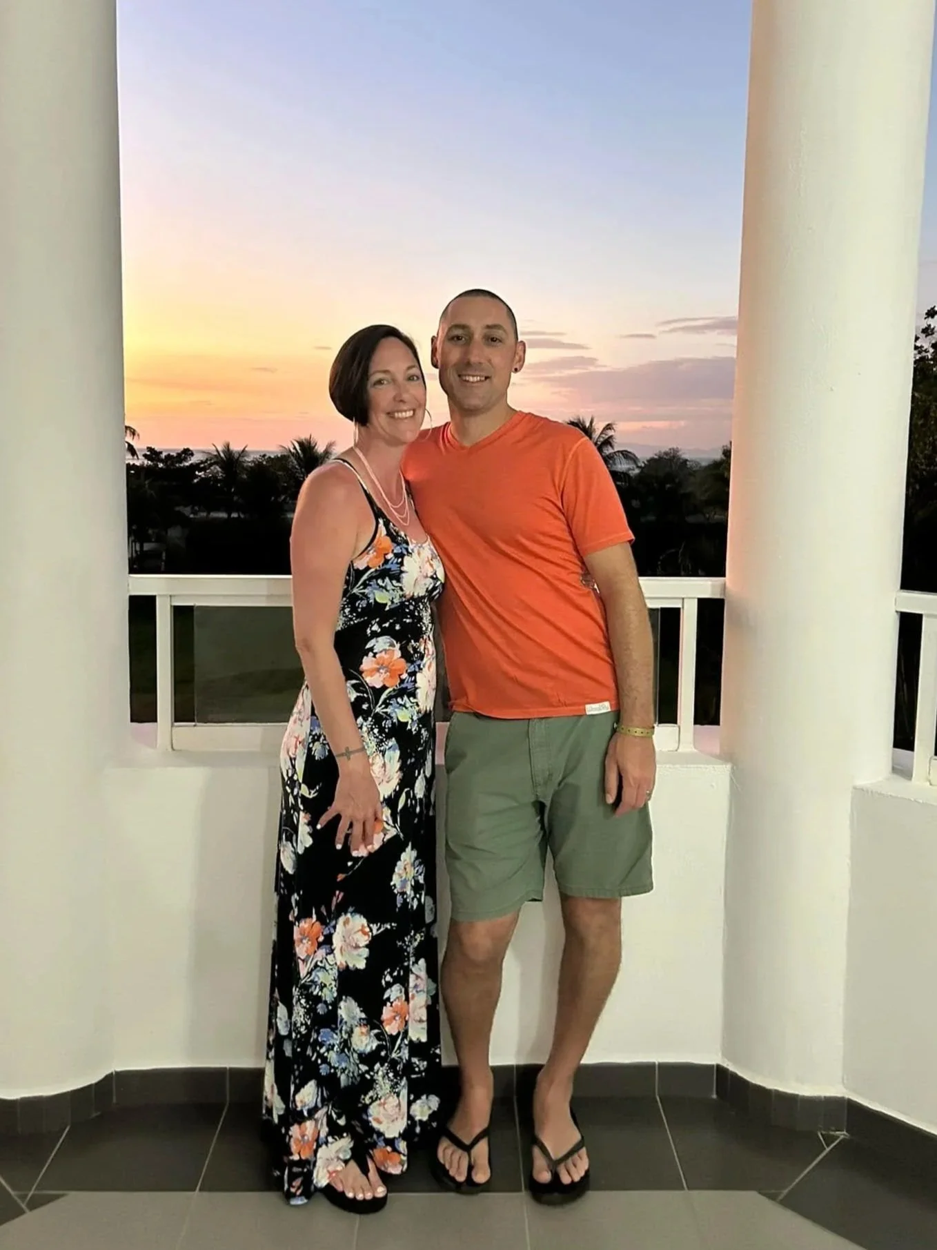 A smiling woman and man standing on a balcony at sunset, with palm trees in the background. The woman is wearing a floral dress and necklace, and the man is dressed in an orange t-shirt and green shorts.