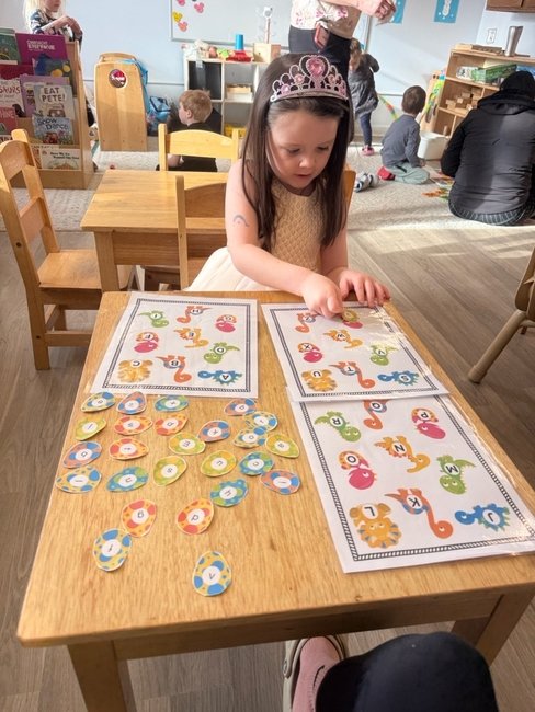 A young girl with a tiara playing a board game with tokens in a classroom setting.
