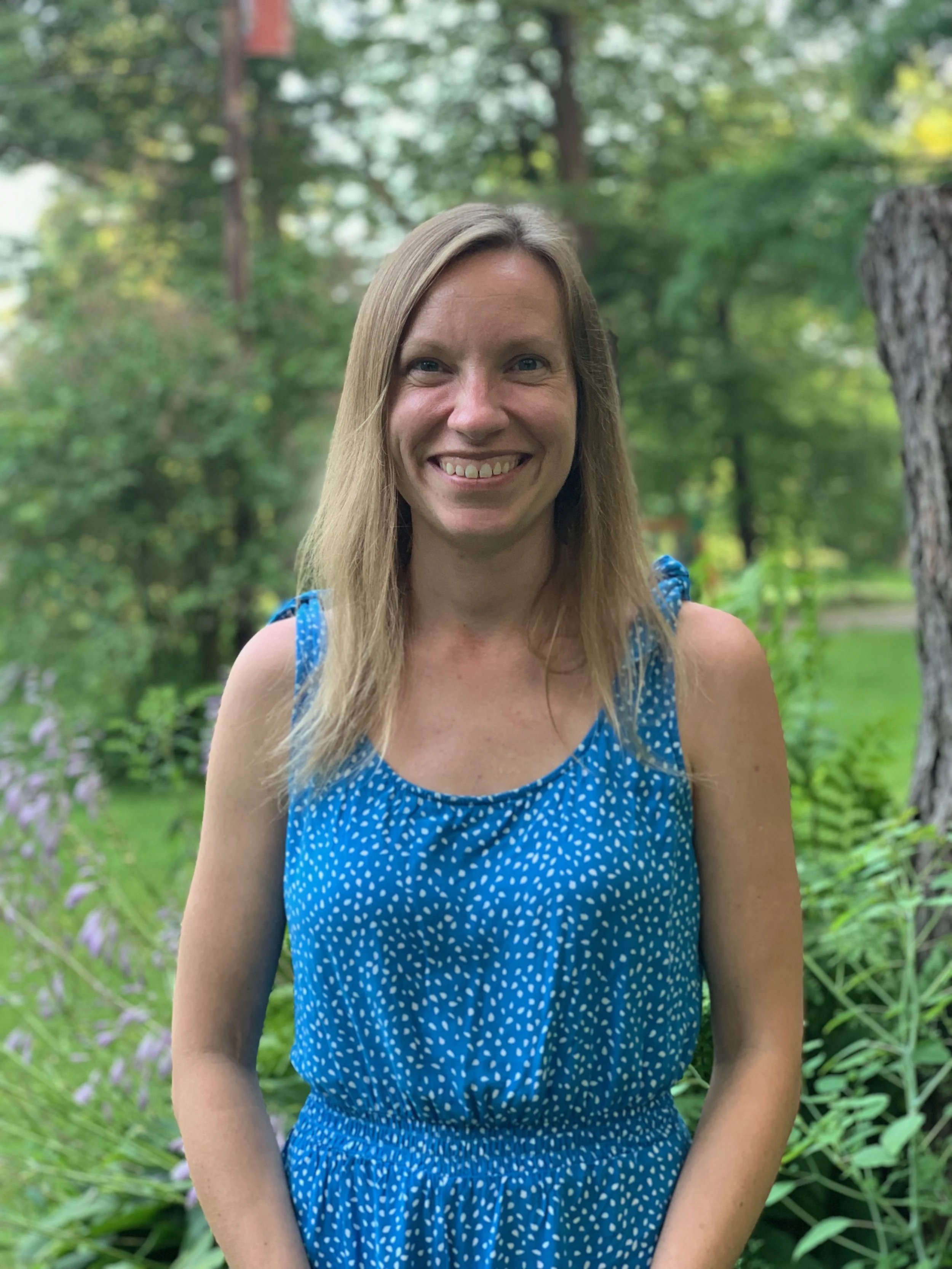 A smiling woman with long blonde hair wearing a blue sleeveless dress standing outdoors in a green garden with trees and plants.