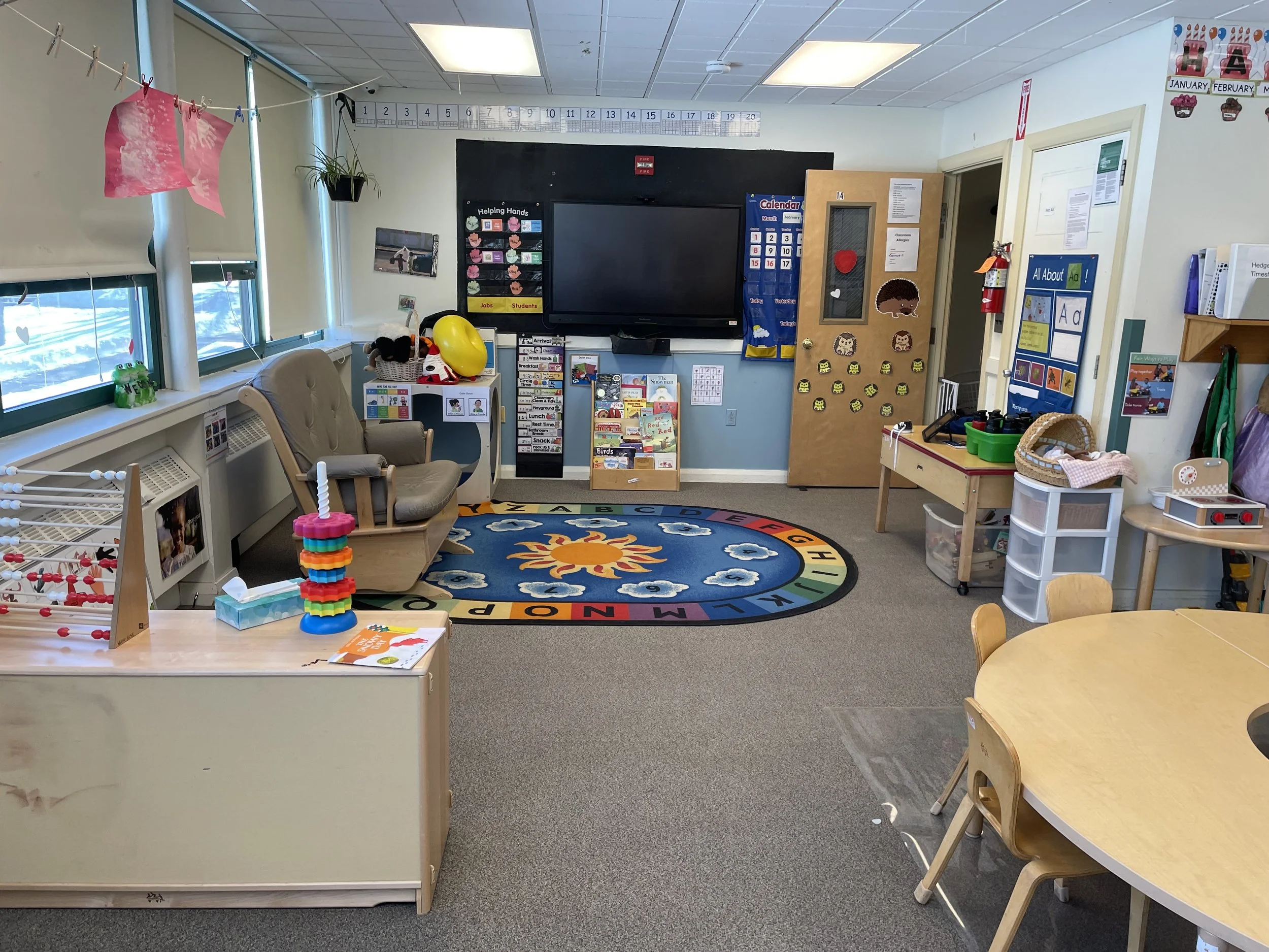 Bright preschool classroom with windows on left, a colorful circular alphabet carpet on beige carpet, small tables and chairs, a cozy armchair, and various educational decorations and toys.