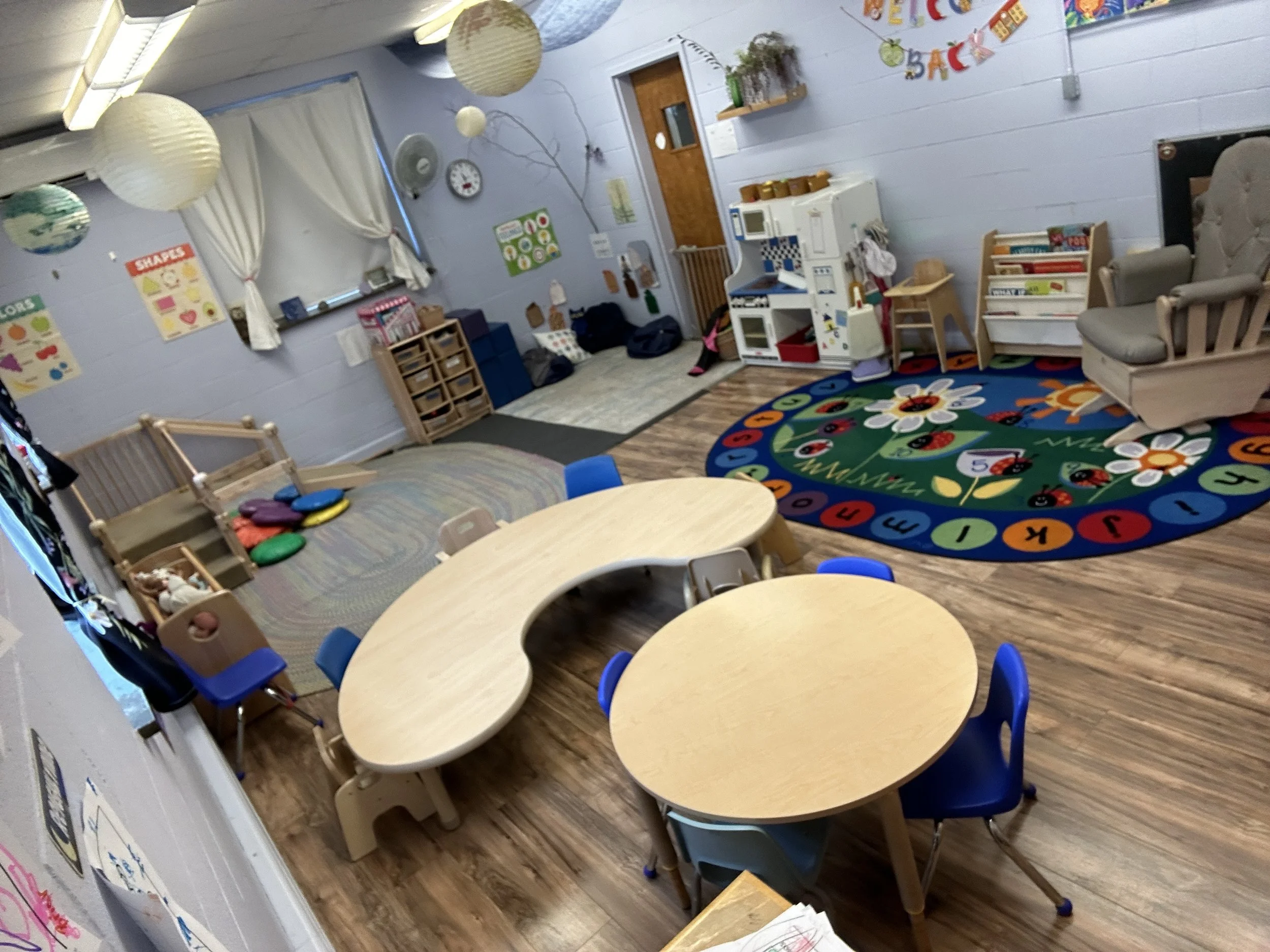A colorful preschool classroom with a wooden floor, round tables with blue and tan chairs, a rainbow-themed rug, a pretend play kitchen, a cozy dollhouse area, and educational posters on the walls.