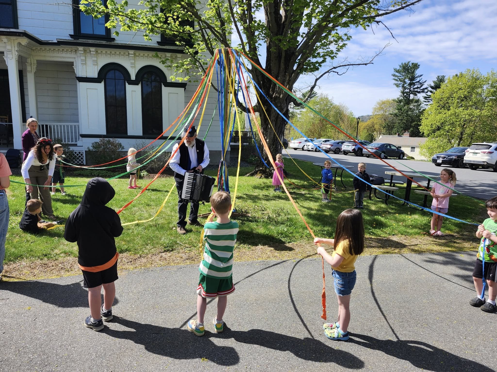 Children and adults gather outdoors in a circle holding colorful ribbons attached to a central person playing an accordion near a large tree, with a building and parked cars in the background.