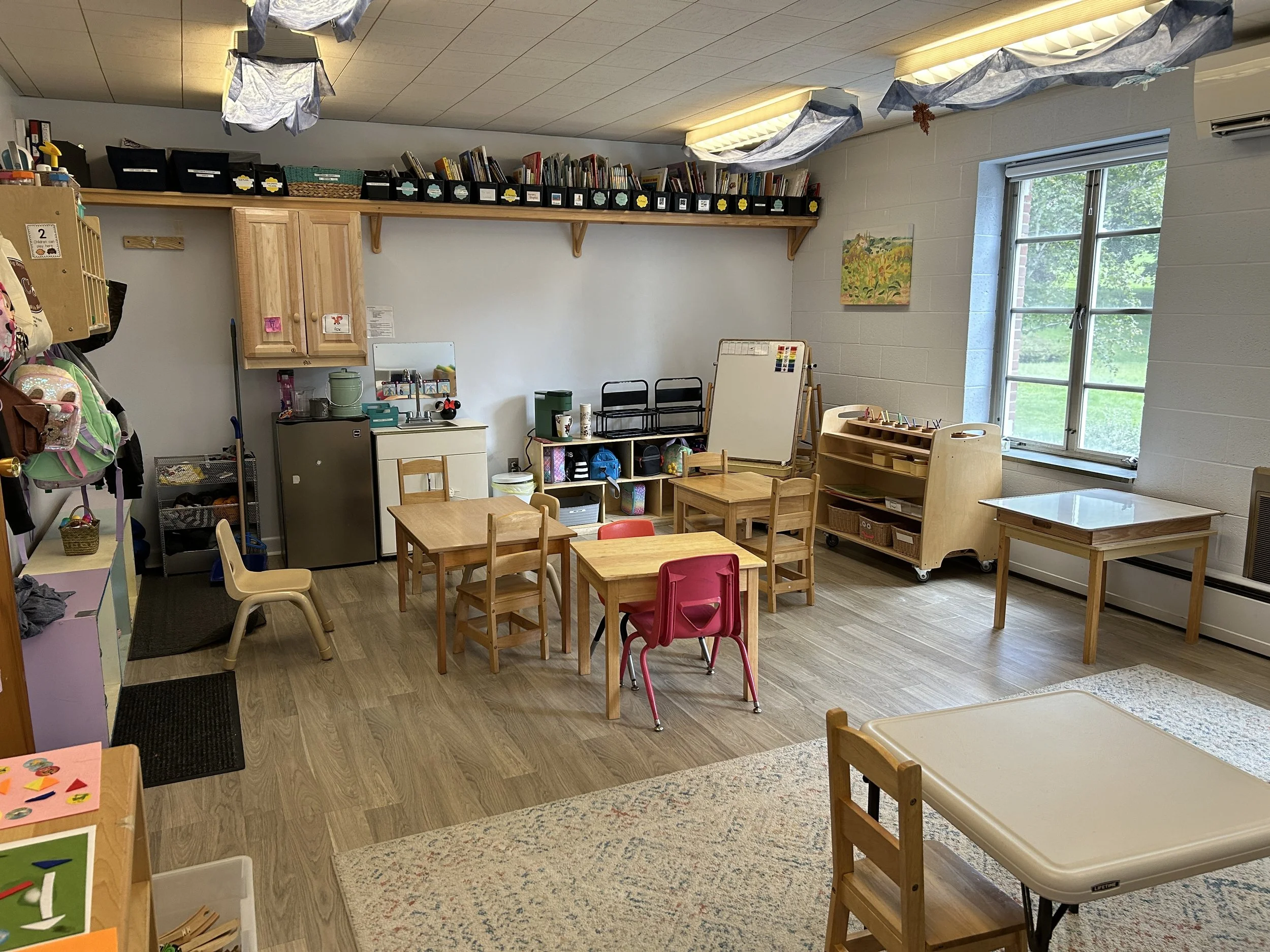 A preschool classroom with small wooden tables and chairs, a window with a view of greenery, storage shelves, and various educational materials.
