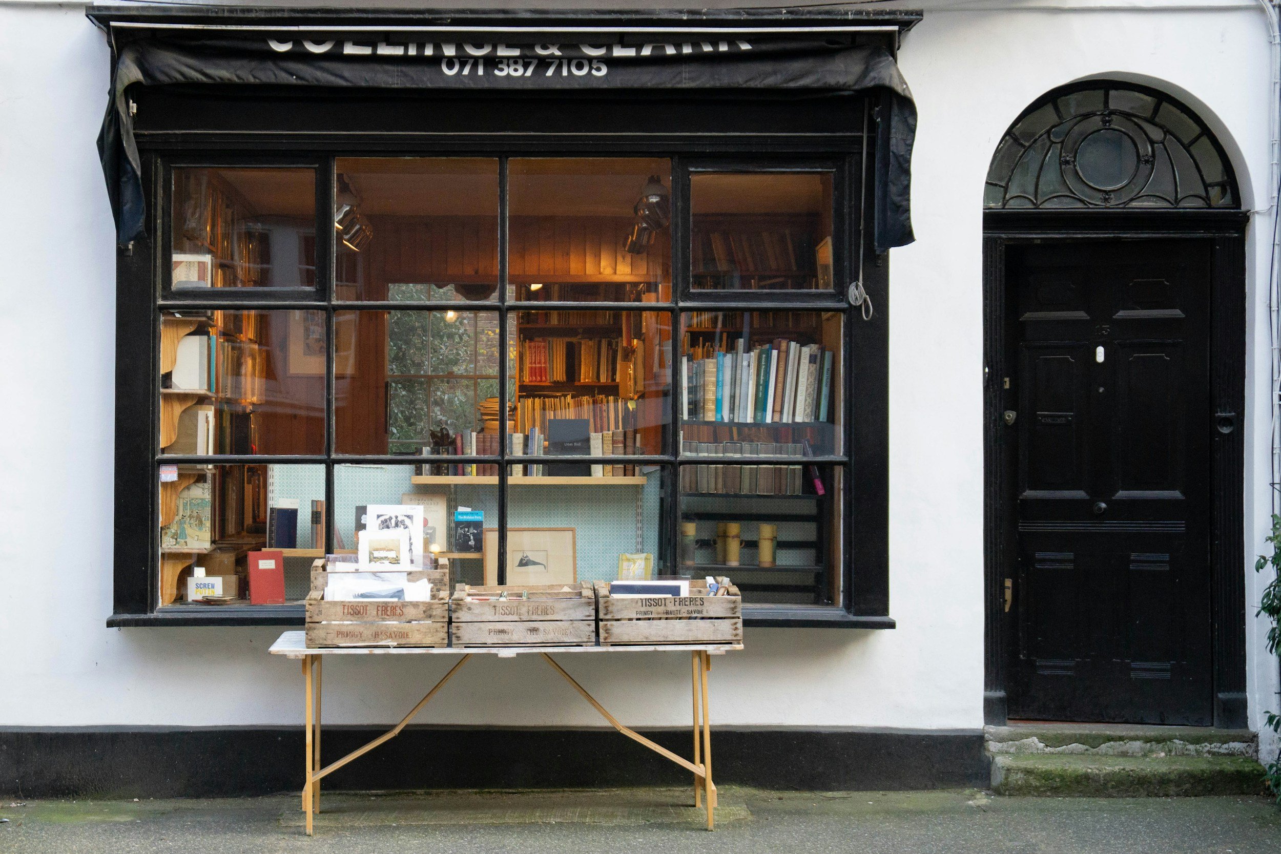 A bookstore with a large window displaying shelves filled with books inside, and a black door next to it. Outside, a small table with wooden crates contains more books and items.