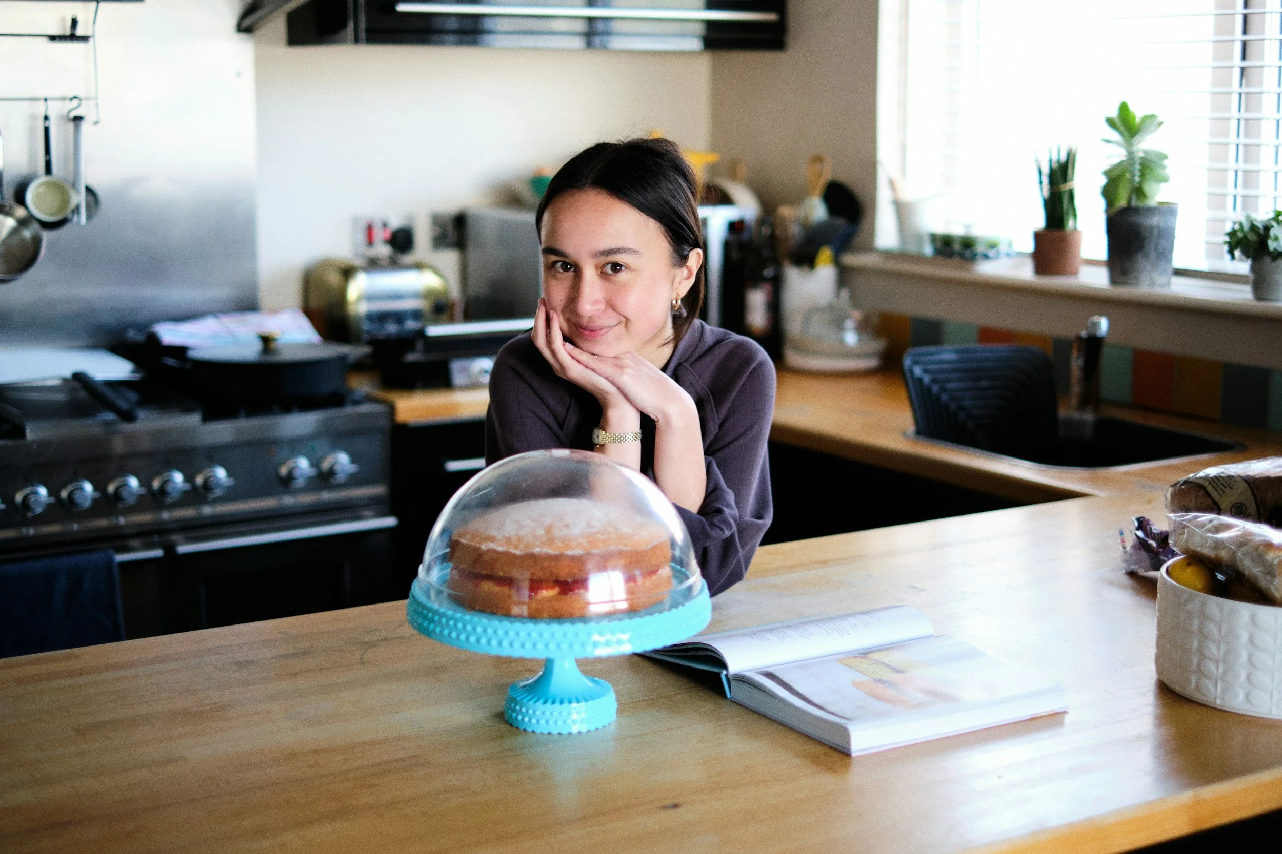 A young woman with dark hair in a kitchen, smiling and resting her chin on her hand, sitting behind a cake on a blue cake stand.
