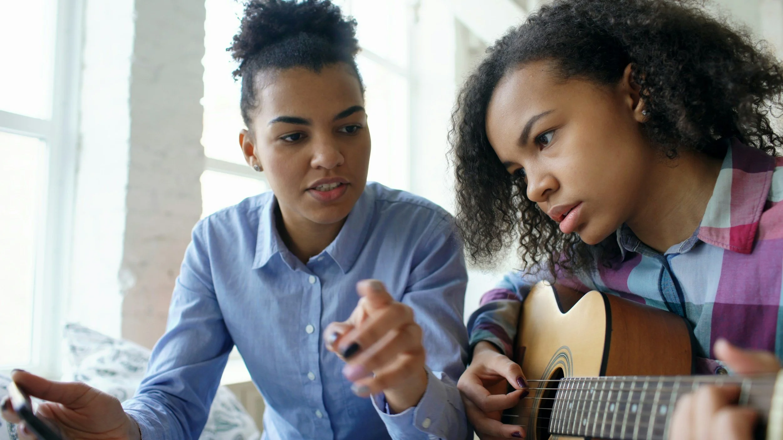 Two women sitting close together, one holding a smartphone and the other playing an acoustic guitar, engaged in a discussion or music lesson in a bright room.