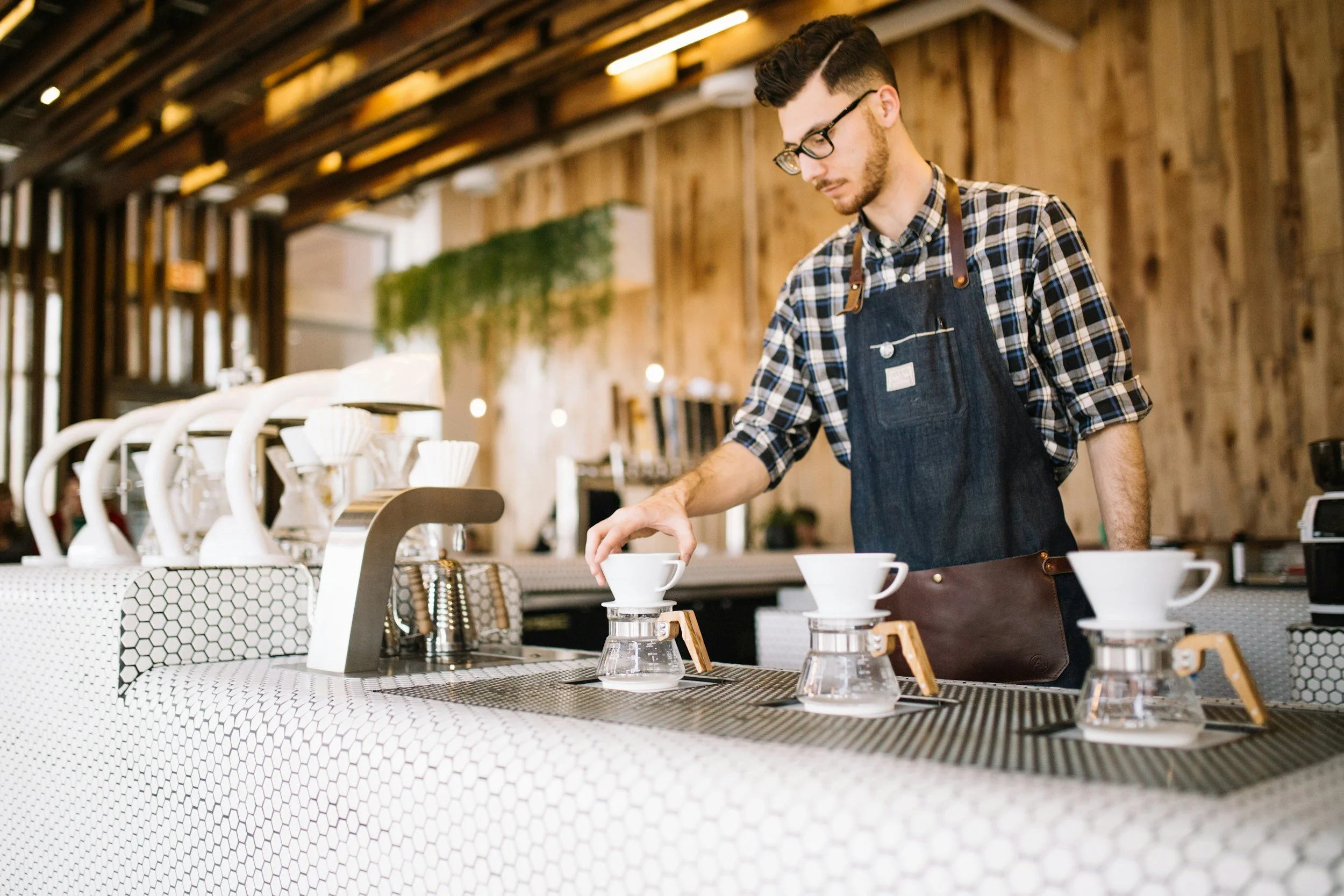Barista preparing pour-over coffee with three drippers and glass cups in a modern coffee shop.