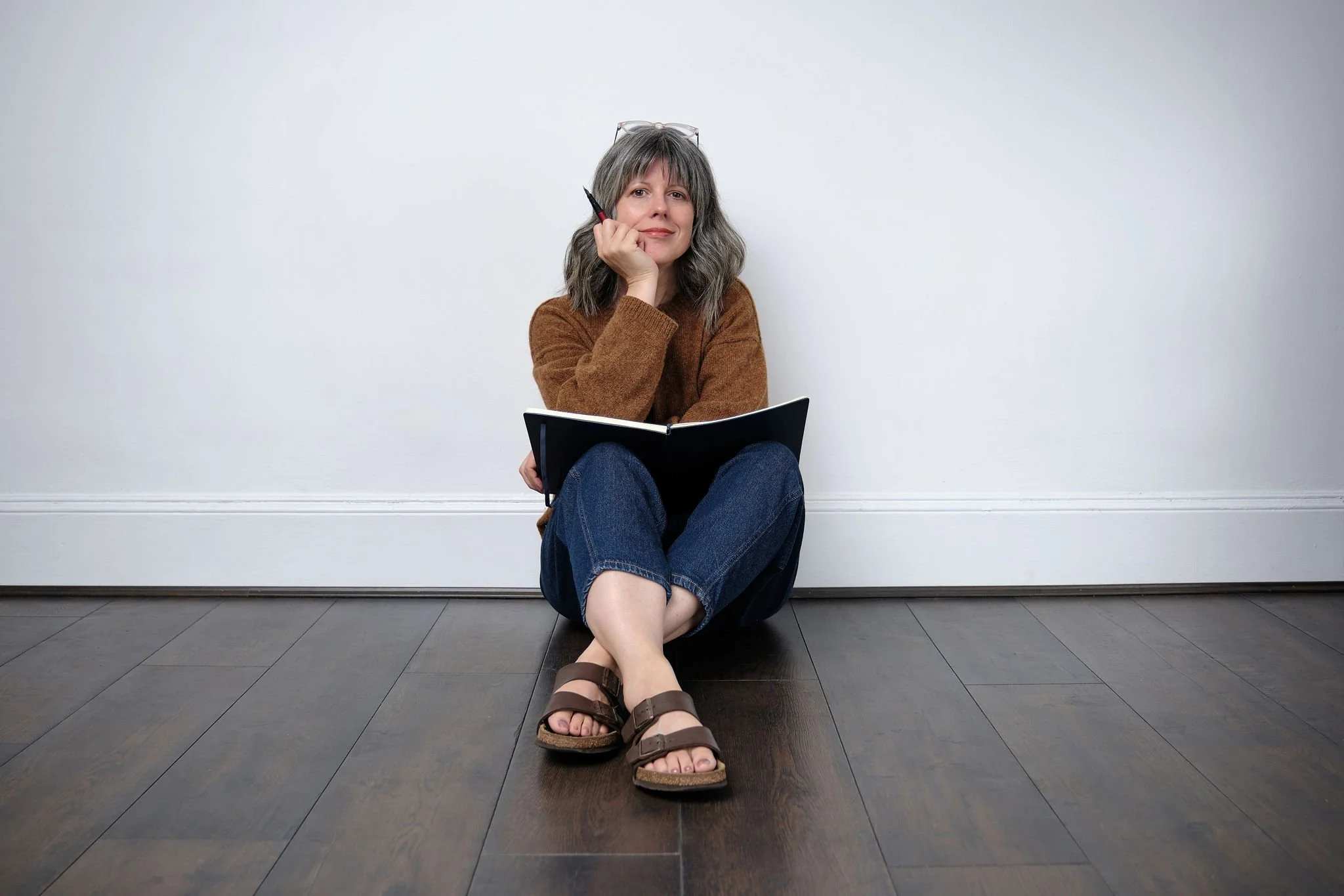 Woman sitting on the floor with crossed legs, holding a notebook and a pen, resting her head on her hand, looking thoughtful.