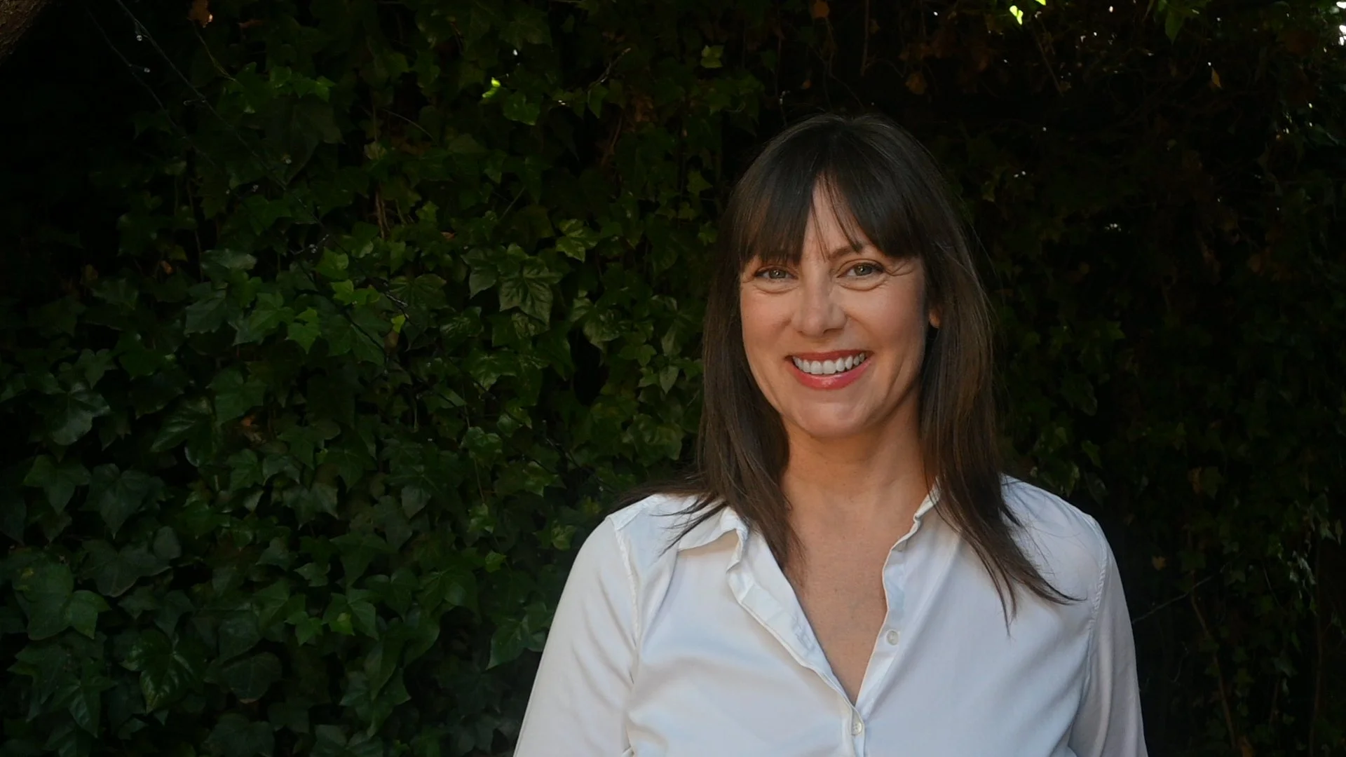 A woman with brown hair and bangs smiling outdoors in front of a dark green leafy background, wearing a white button-up shirt.