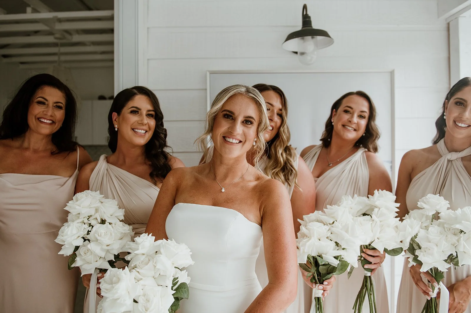 A bride and five bridesmaids smiling, holding white bouquets, indoors with white wooden wall and black ceiling light.