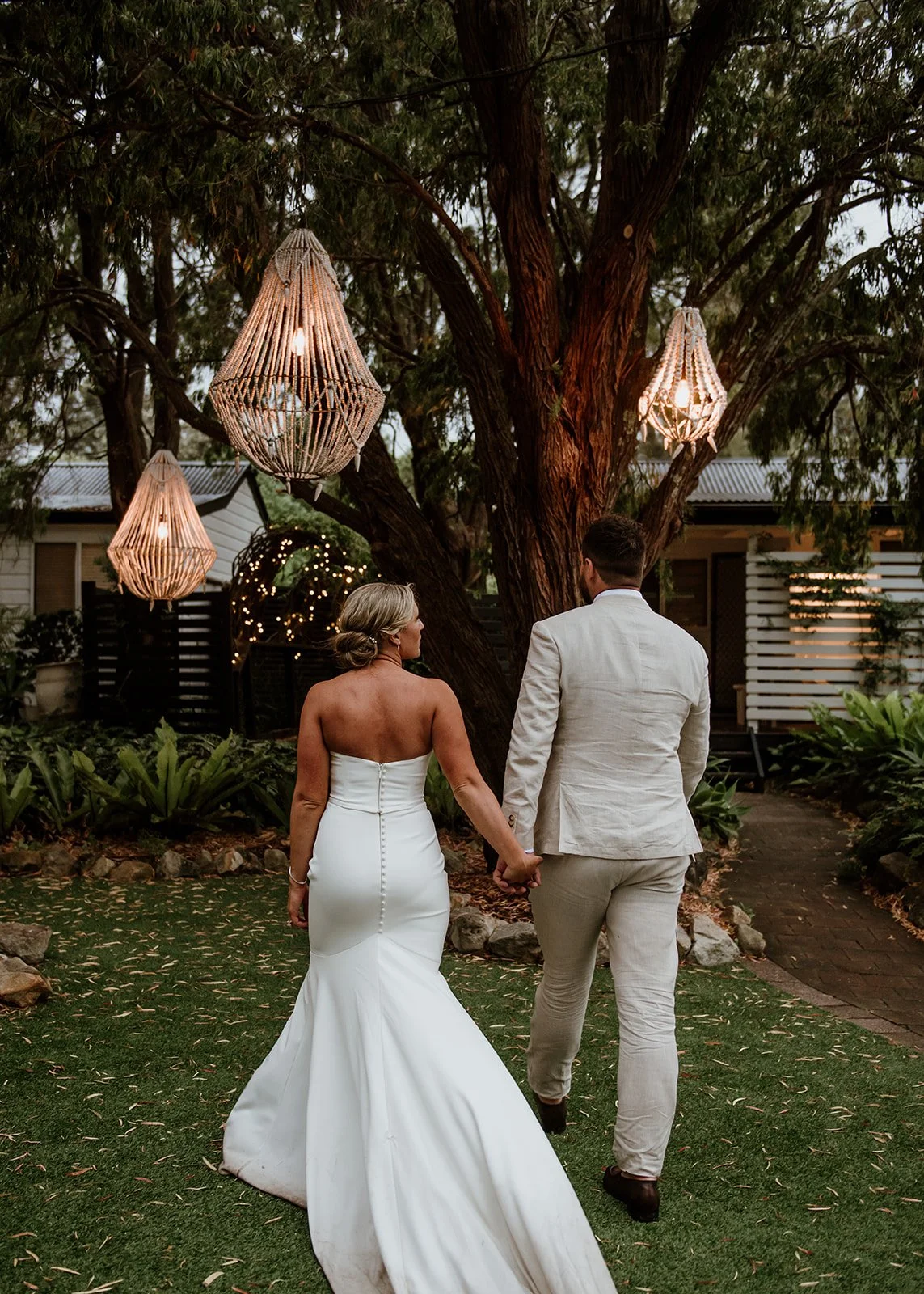 A bride and groom walking hand in hand outdoors during their wedding, surrounded by trees and hanging lanterns at dusk.