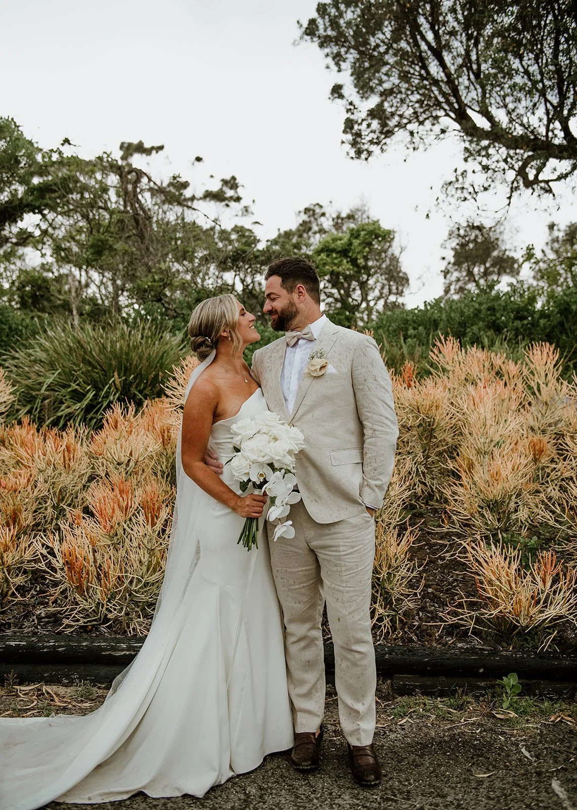 A bride and groom in wedding attire stand close together outdoors, smiling and gazing into each other's eyes, with desert plants and trees in the background.