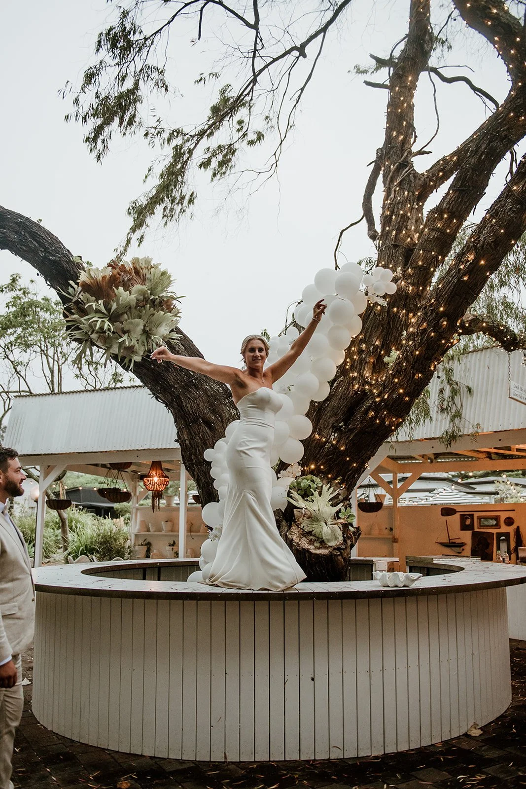 Bride in a white wedding dress standing on a raised platform around a large tree decorated with white balloons and fairy lights, holding a bouquet, with a man standing nearby and a garden setting in the background.