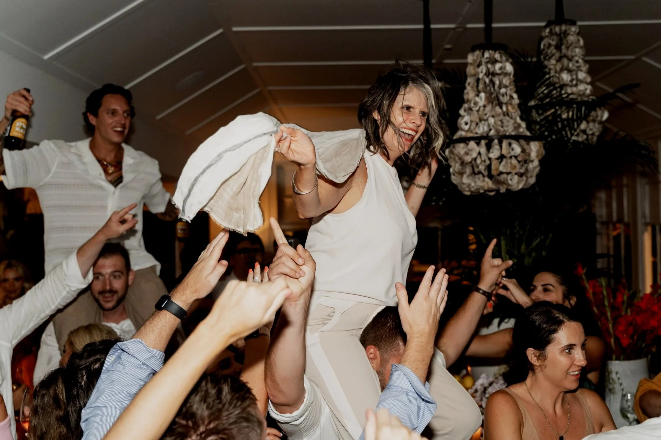 People celebrating at a party with one woman being lifted on a man's shoulders, holding a towel or cloth, smiling, and surrounded by others dancing and cheering.