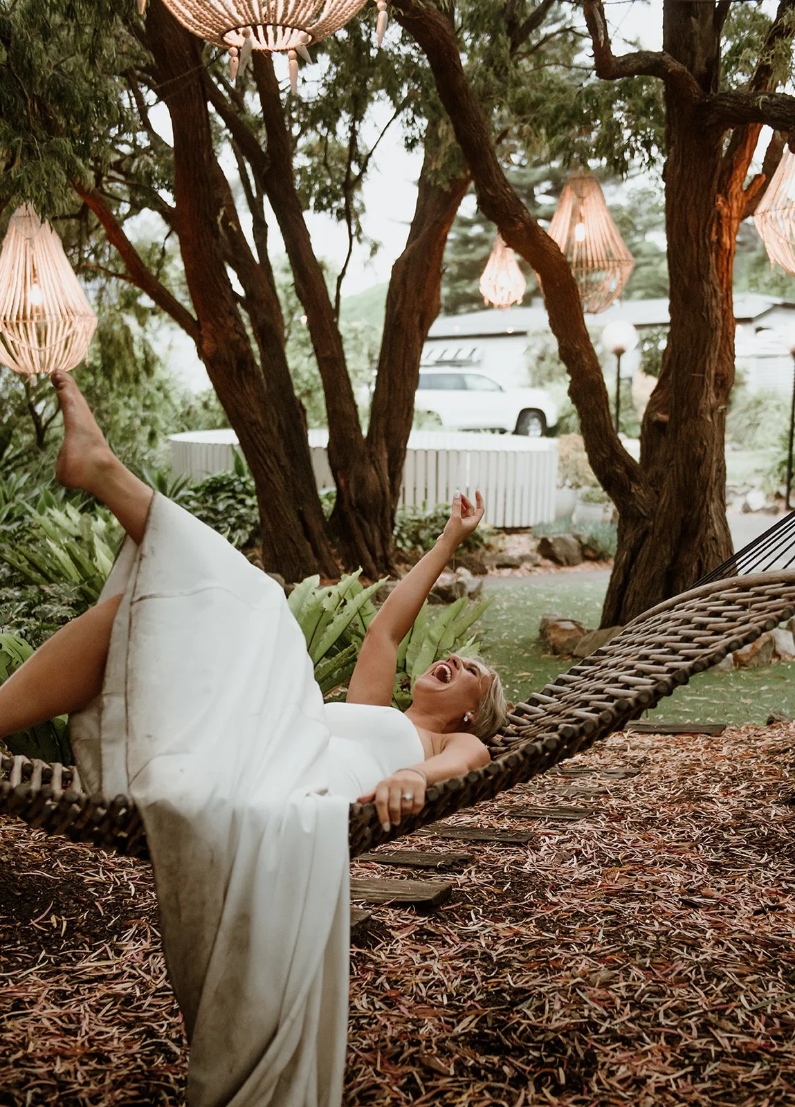 A woman in a white dress is lying and laughing on a hammock hanging between trees in a backyard, with decorative hanging lanterns above her.