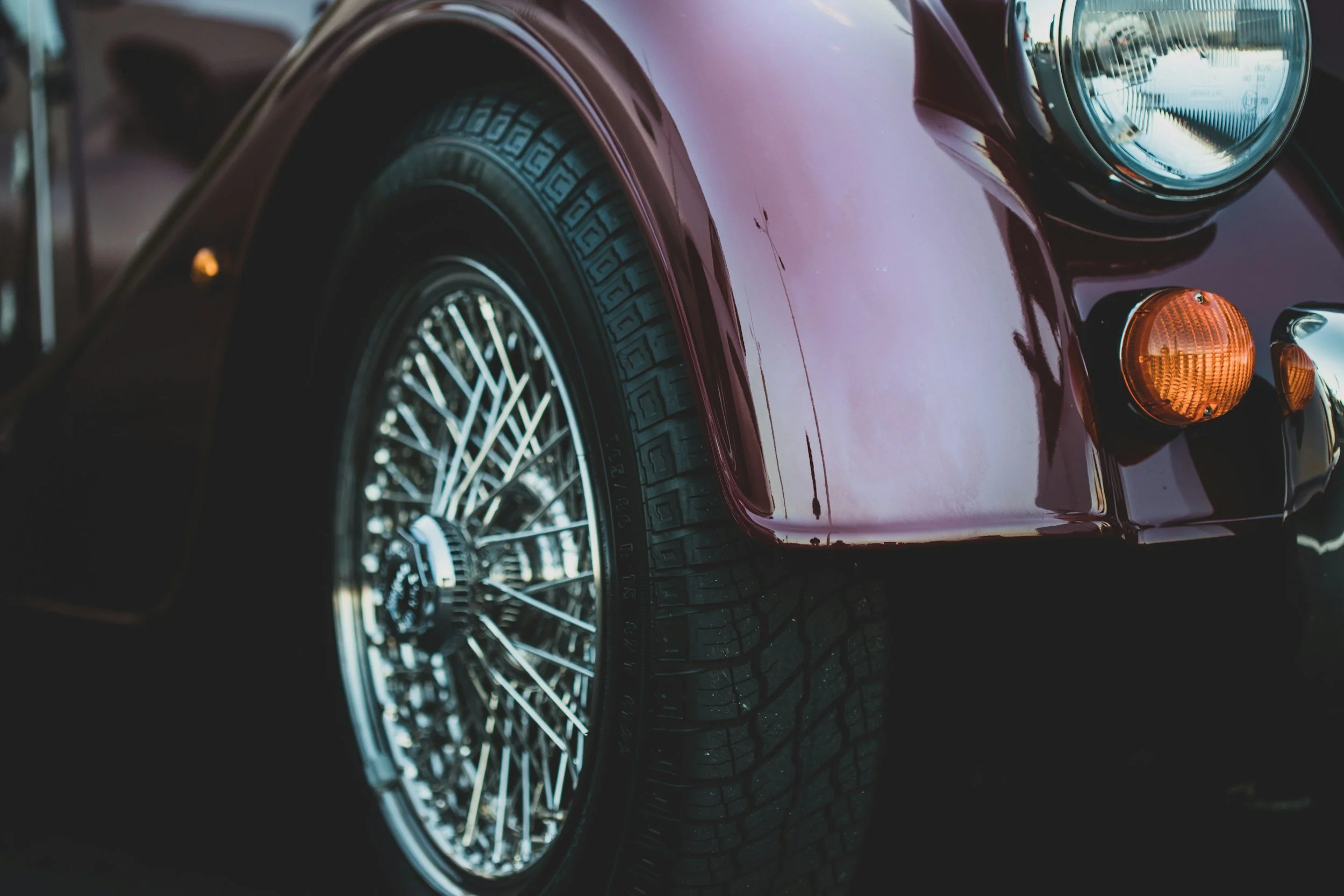 Close-up of the front end of a vintage purple sports car, showing the headlight, turn signal, and wire-spoked wheel.
